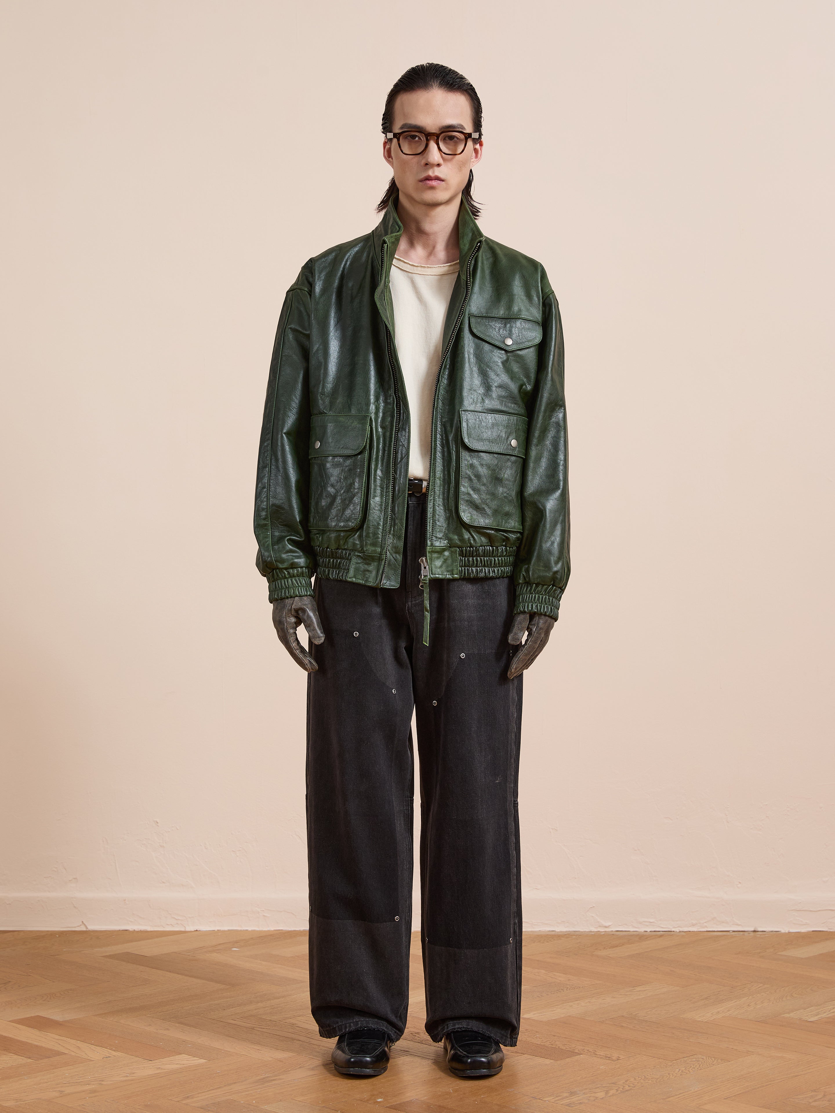 A person stands against a plain wall wearing the FOUND Forest Leather Bomber Jacket, paired with a white shirt, wide black pants, black shoes, gloves, and glasses.