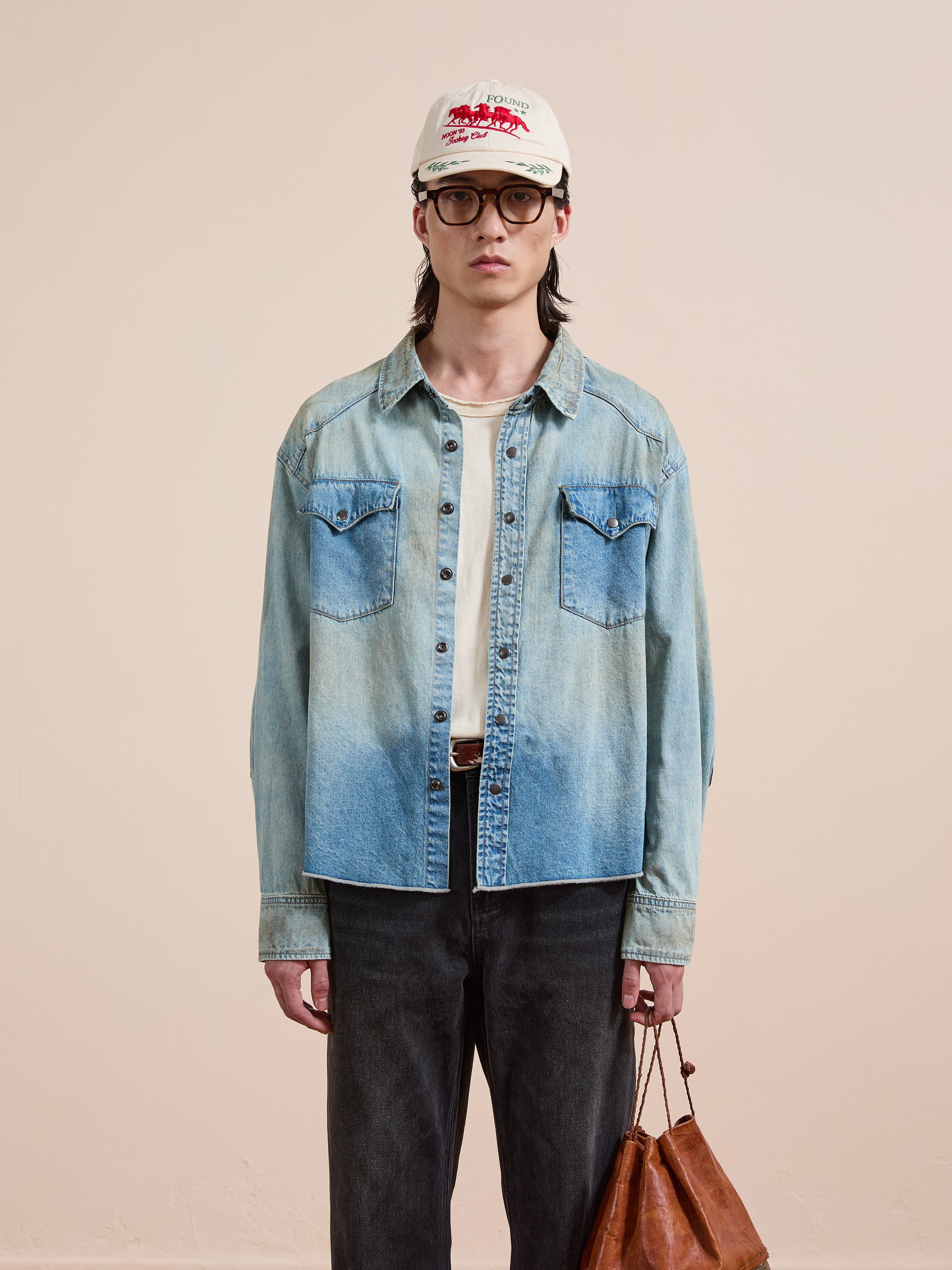 Wearing the FOUND Denim Western Snap Button Shirt, black jeans, glasses, and a white cap, a person stands against a plain background holding a brown leather bag.