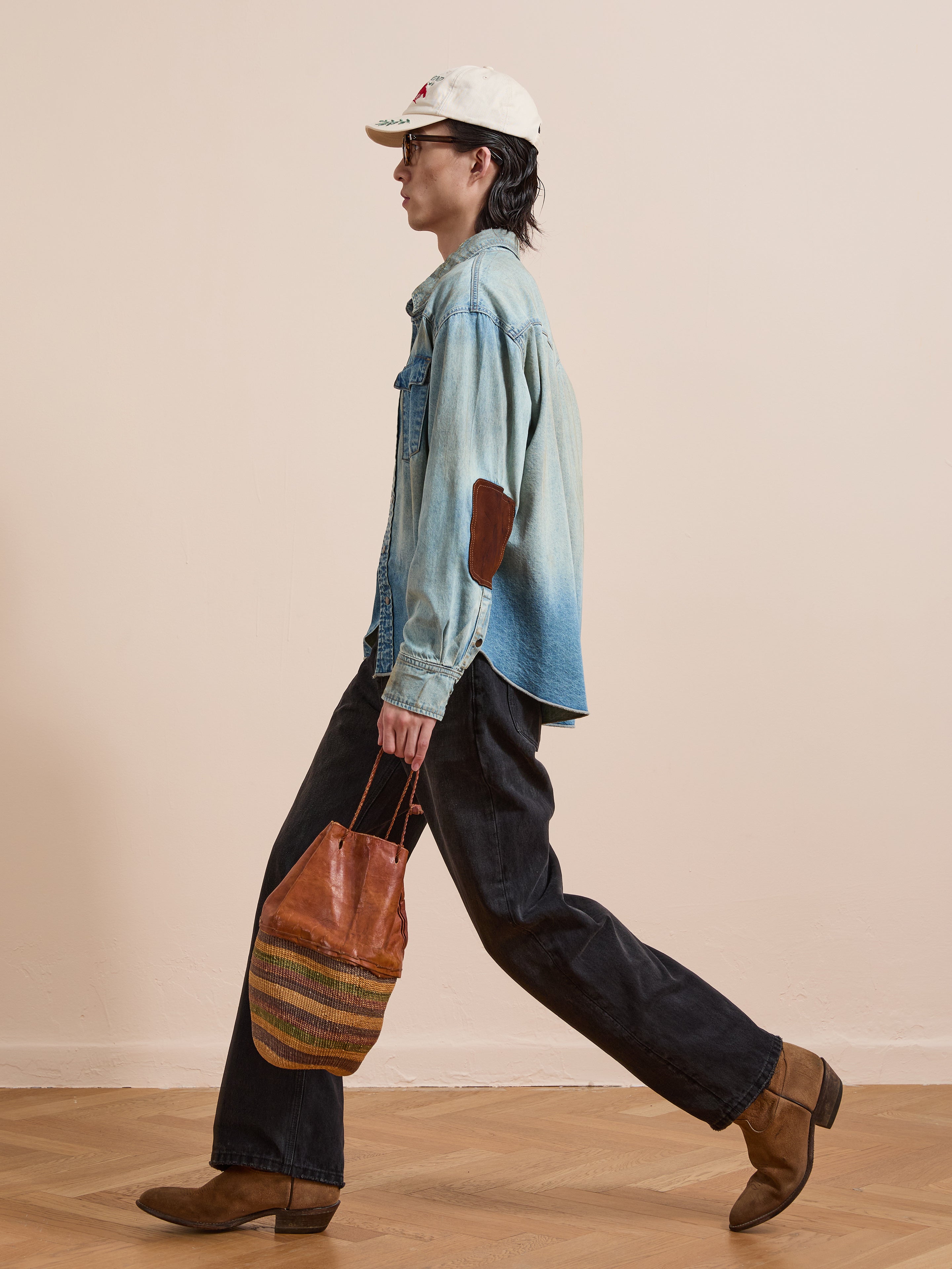 A person walks indoors wearing a white cap, sunglasses, the FOUND Denim Western Snap Button Shirt with vintage-wash, dark jeans, brown boots, and carrying a brown striped bag.