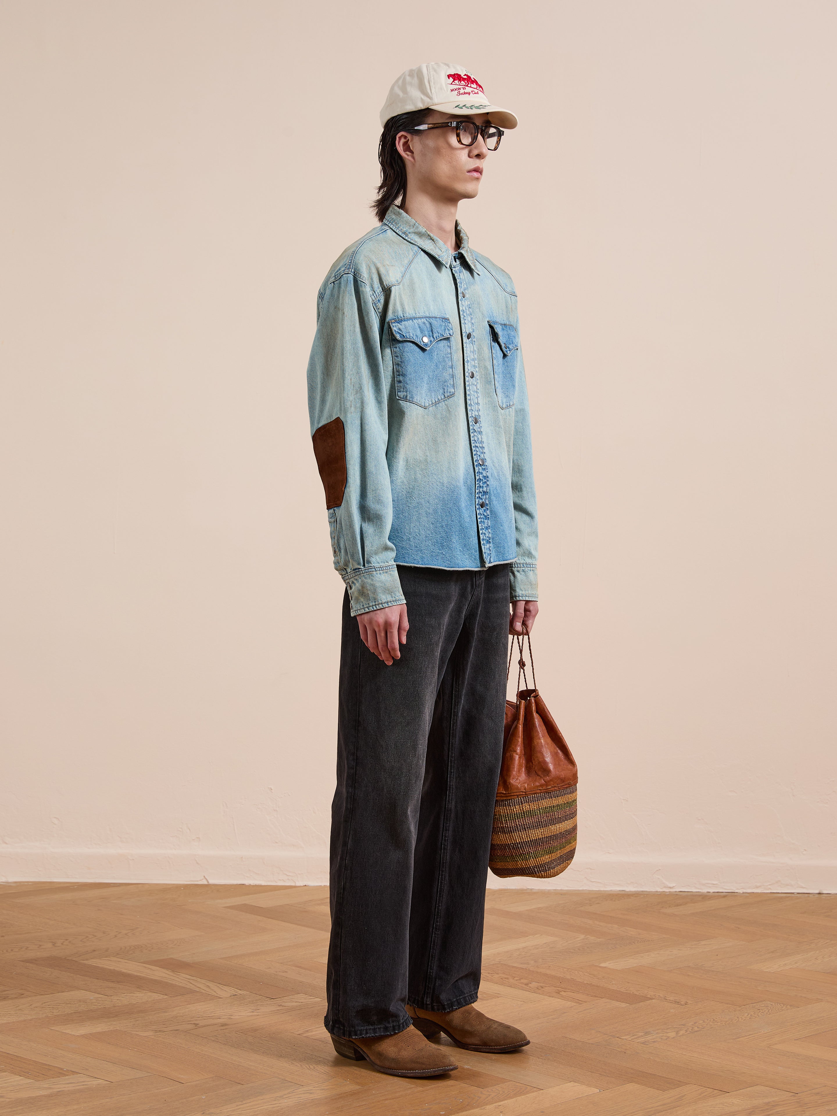 Indoors, a person wears the FOUND Denim Western Snap Button Shirt with western yoke detailing, paired with black jeans, brown shoes, a white baseball cap, glasses, and holds a patterned brown bag.