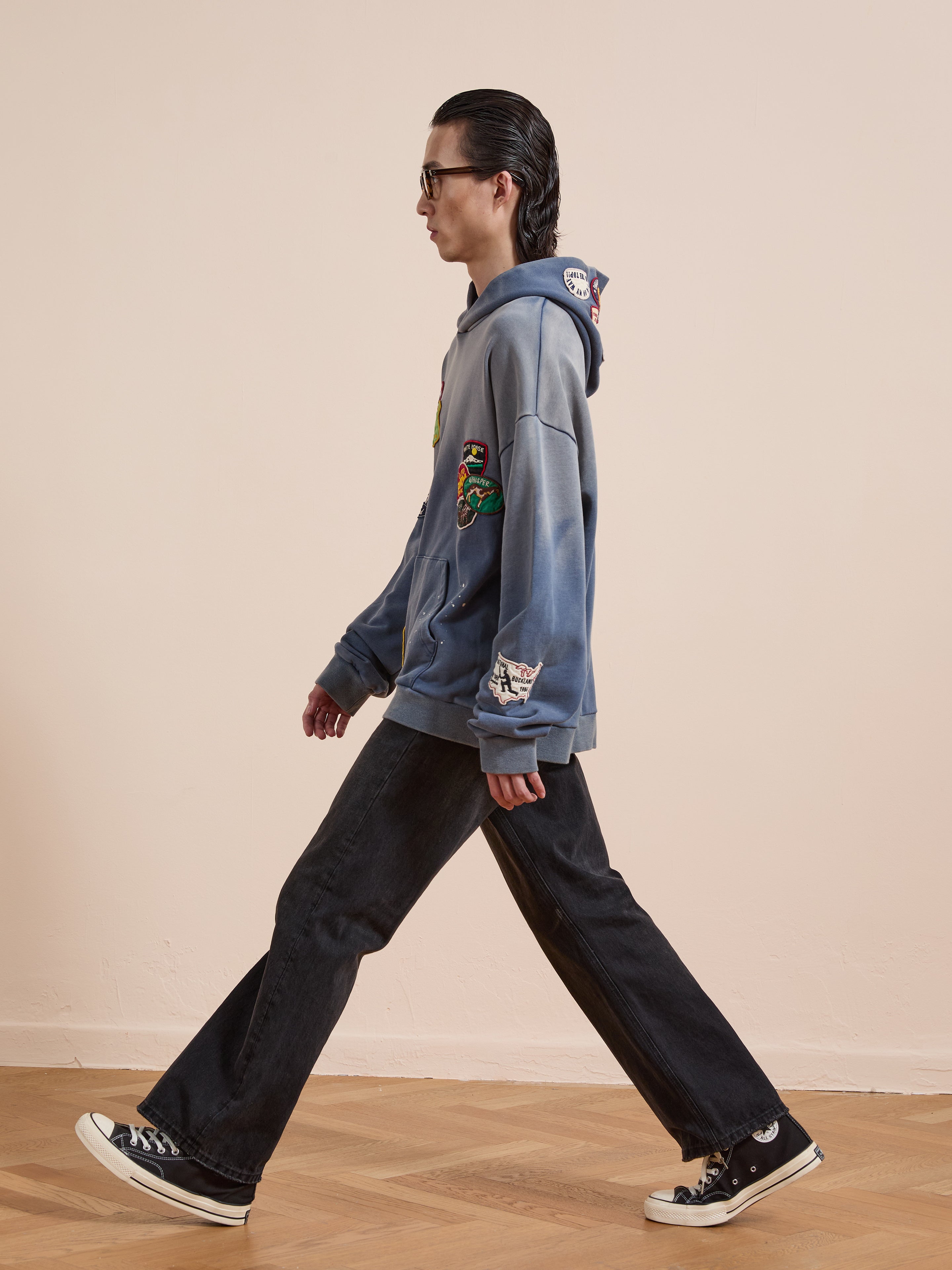 A person with long hair walks indoors, wearing glasses, a blue FOUND Distressed Patch Hoodie with graphic design, black jeans, and black Converse sneakers.