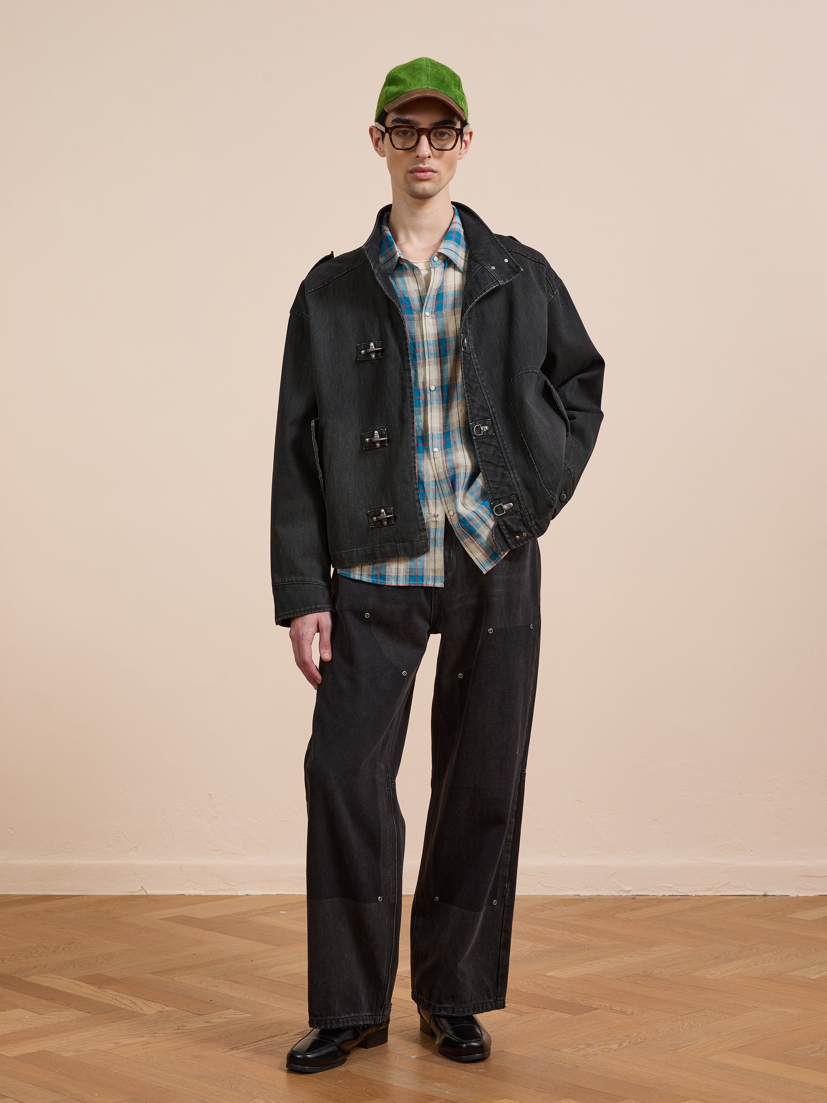 A person wearing a green cap, glasses, wide-leg black pants, black shoes, and the Found Utility Clasp Work Jacket stands on a hardwood floor against a beige wall.
