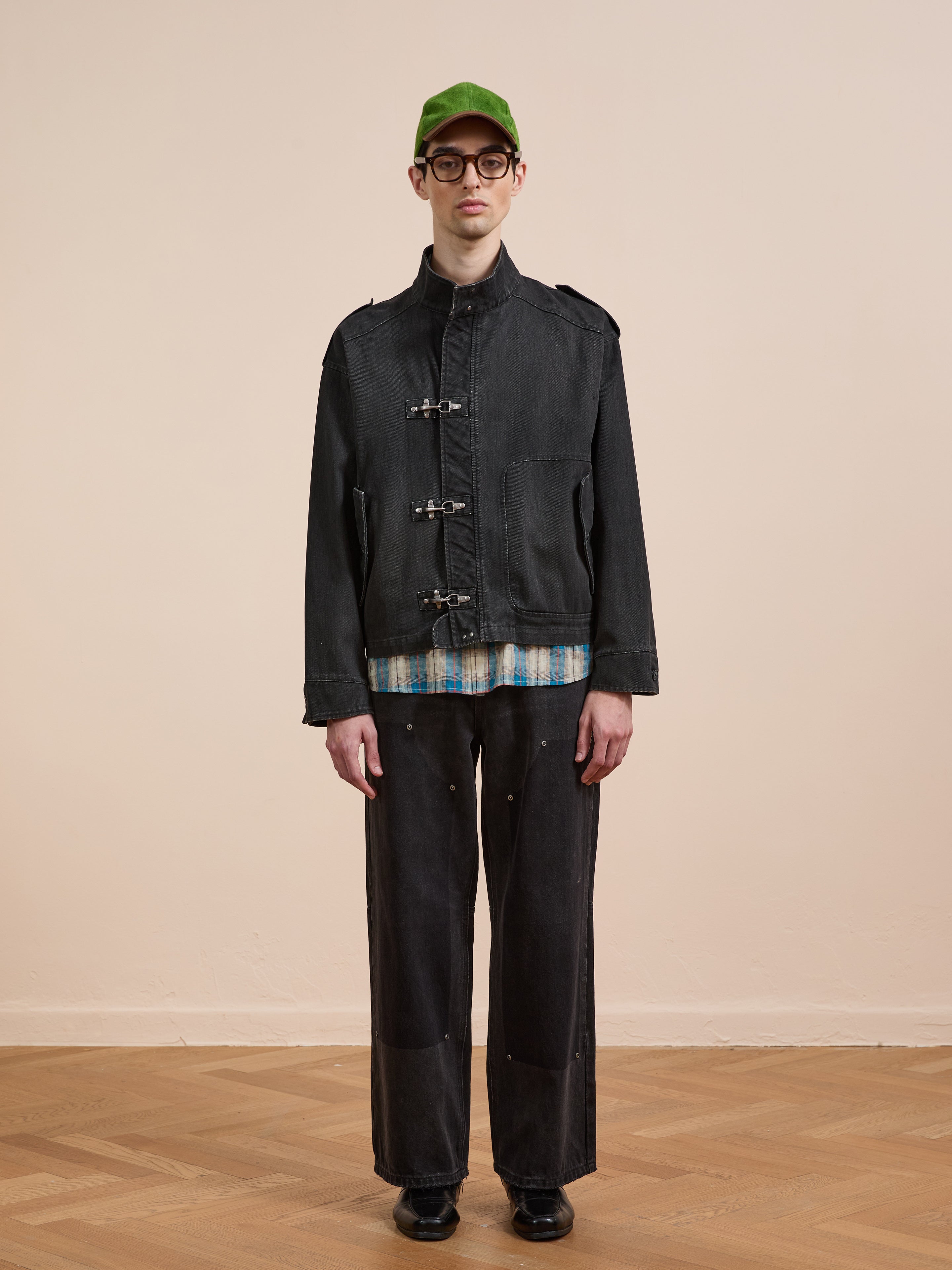 A person in a green cap, glasses, Found Utility Clasp Work Jacket (vintage black), plaid shirt, and wide black pants stands on a wooden floor against a plain beige background.