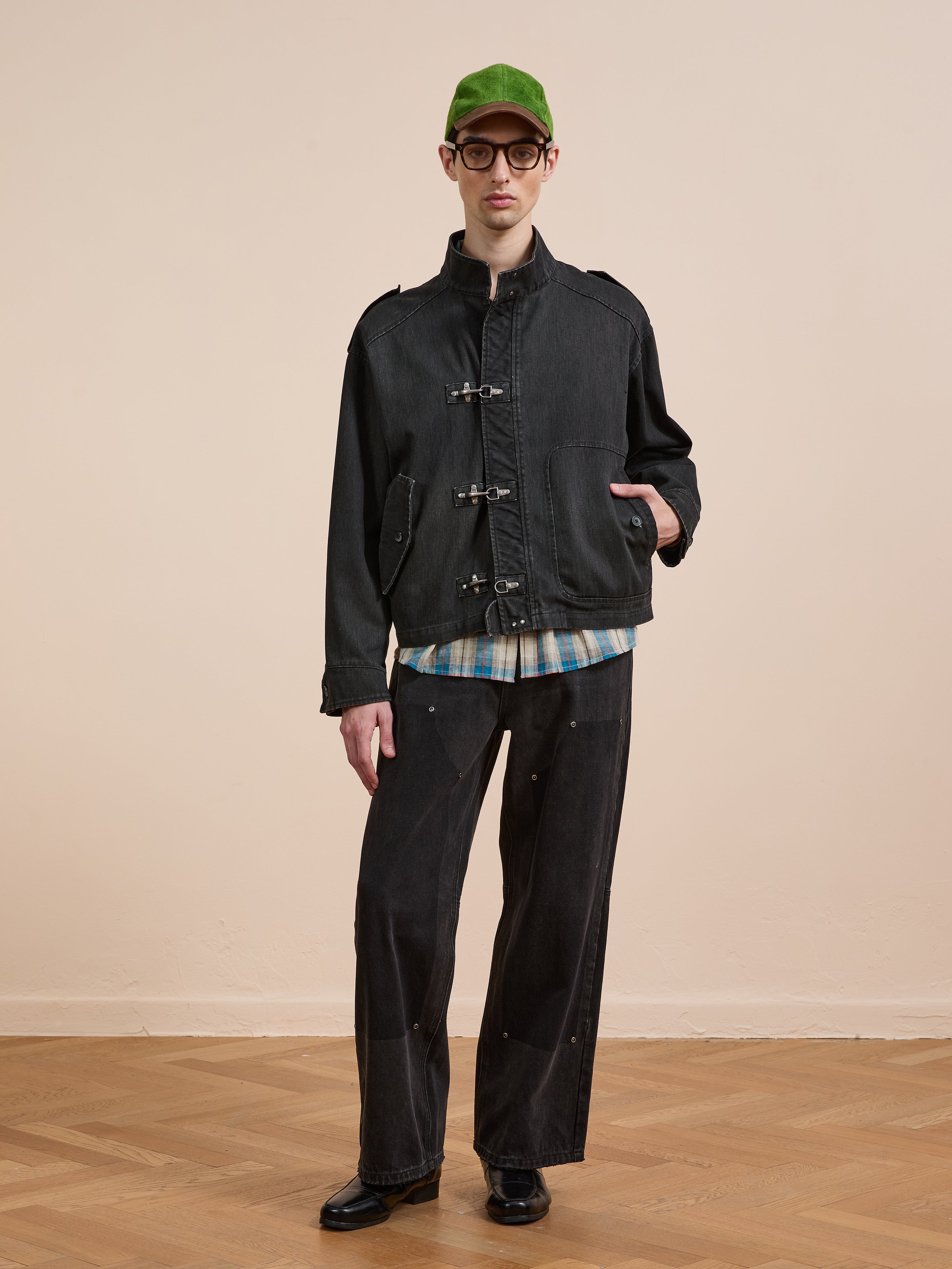 Indoors, a person wears a green cap, round glasses, a plaid shirt, black wide-leg pants and shoes, and the Found Utility Clasp Work Jacket—a vintage-inspired black cotton layer with toggle closures—standing on a wooden floor.