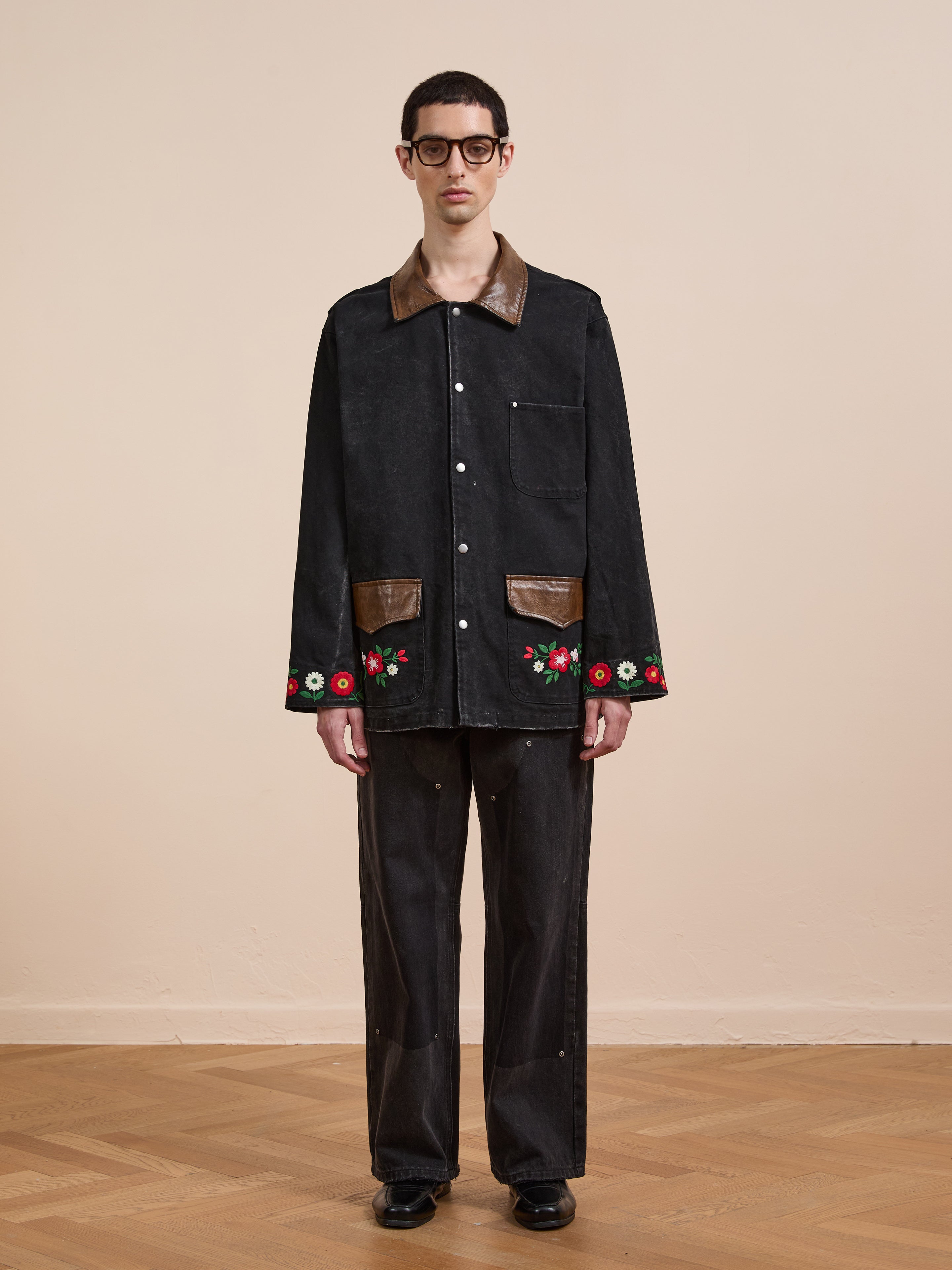 A man in black glasses, wide black pants, and shoes wears the Found Canvas Embroidered Work Jacket— a relaxed-fit unisex black canvas jacket with brown leather accents and floral embroidery—standing by a beige wall on wooden flooring.