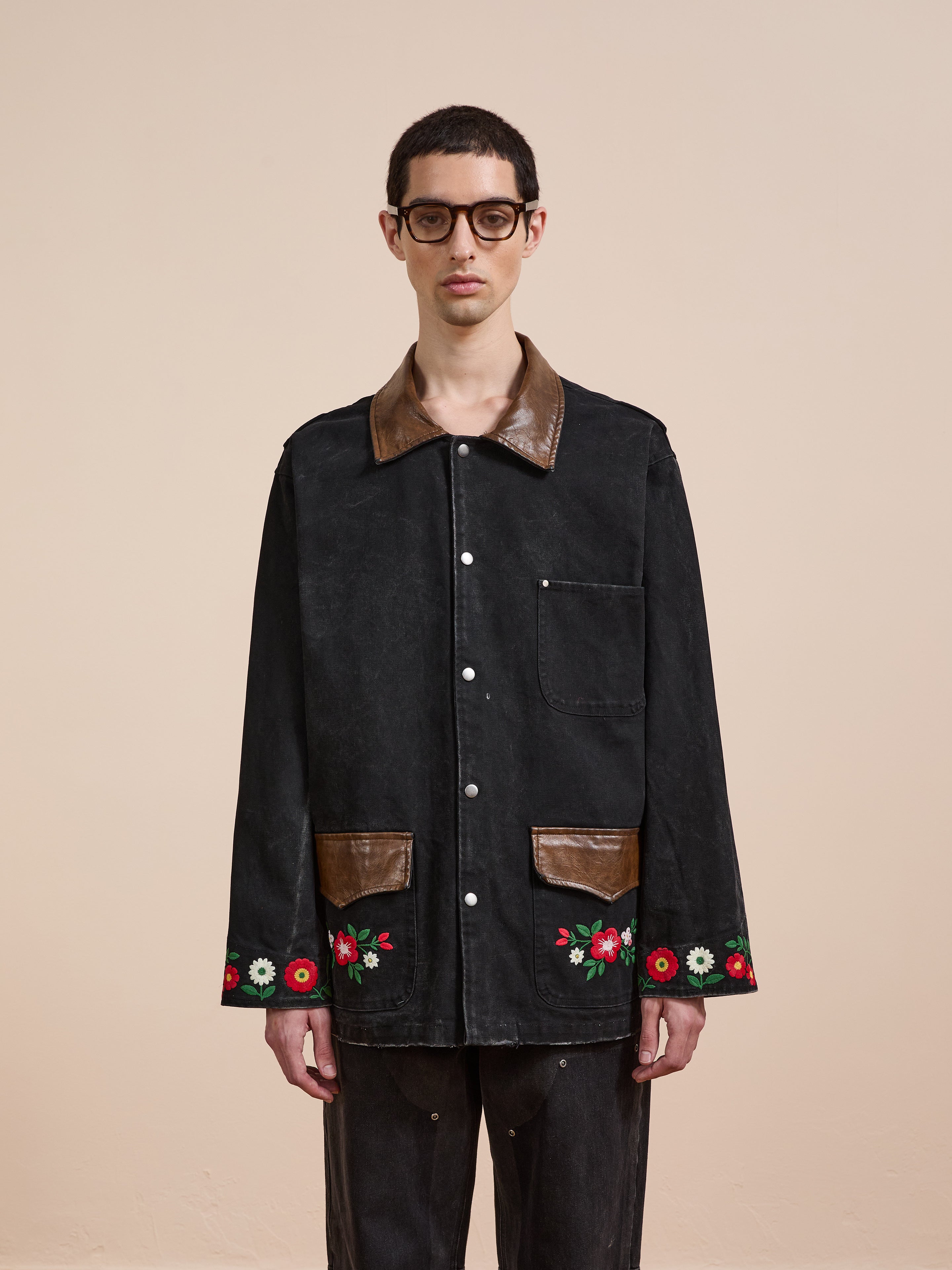 A person wearing glasses and the Found Canvas Embroidered Work Jacket, a unisex relaxed fit black canvas piece with brown leather accents and red floral embroidery, stands against a plain beige background.