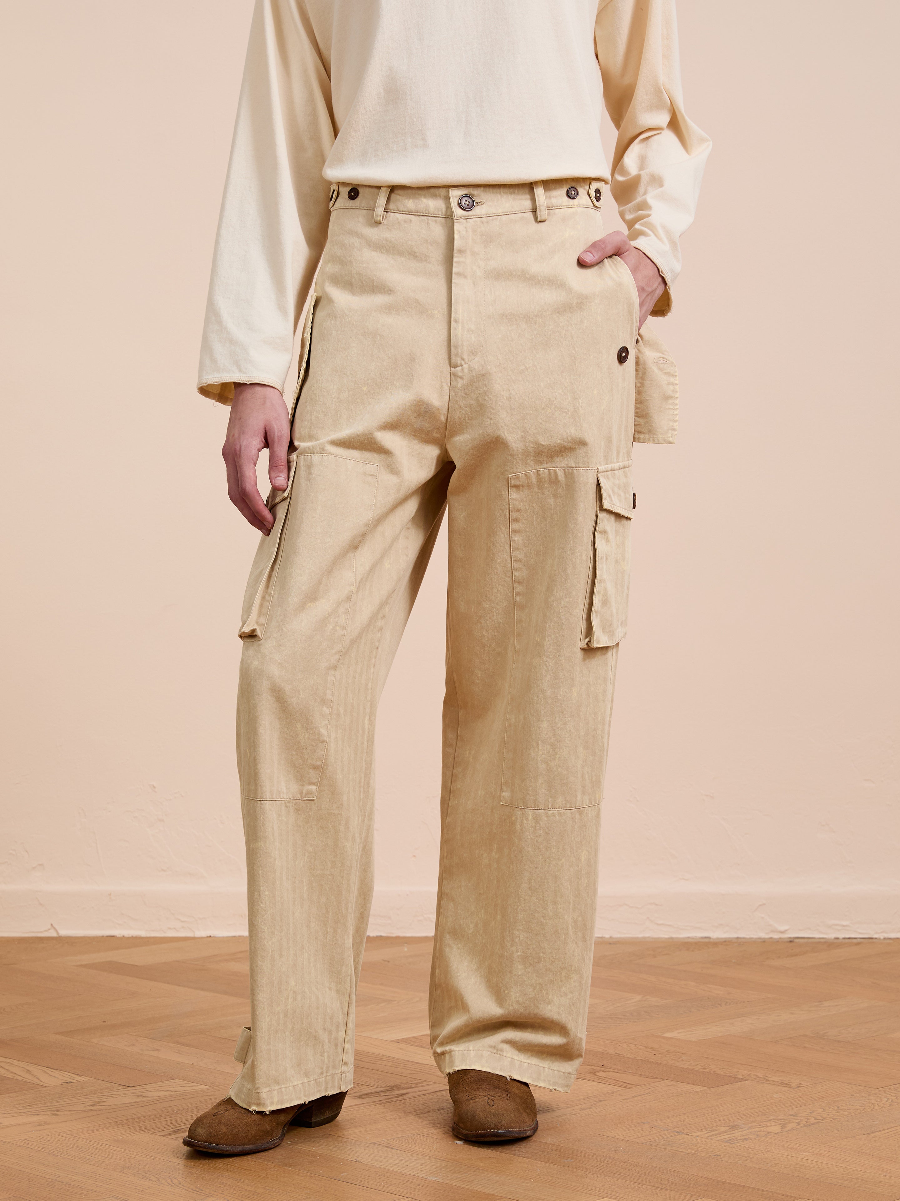 A person in FOUND Utility Herringbone Paneled Pants and a cream long-sleeve shirt stands on a wooden floor against a neutral backdrop, highlighting the brand’s effortless utility style.