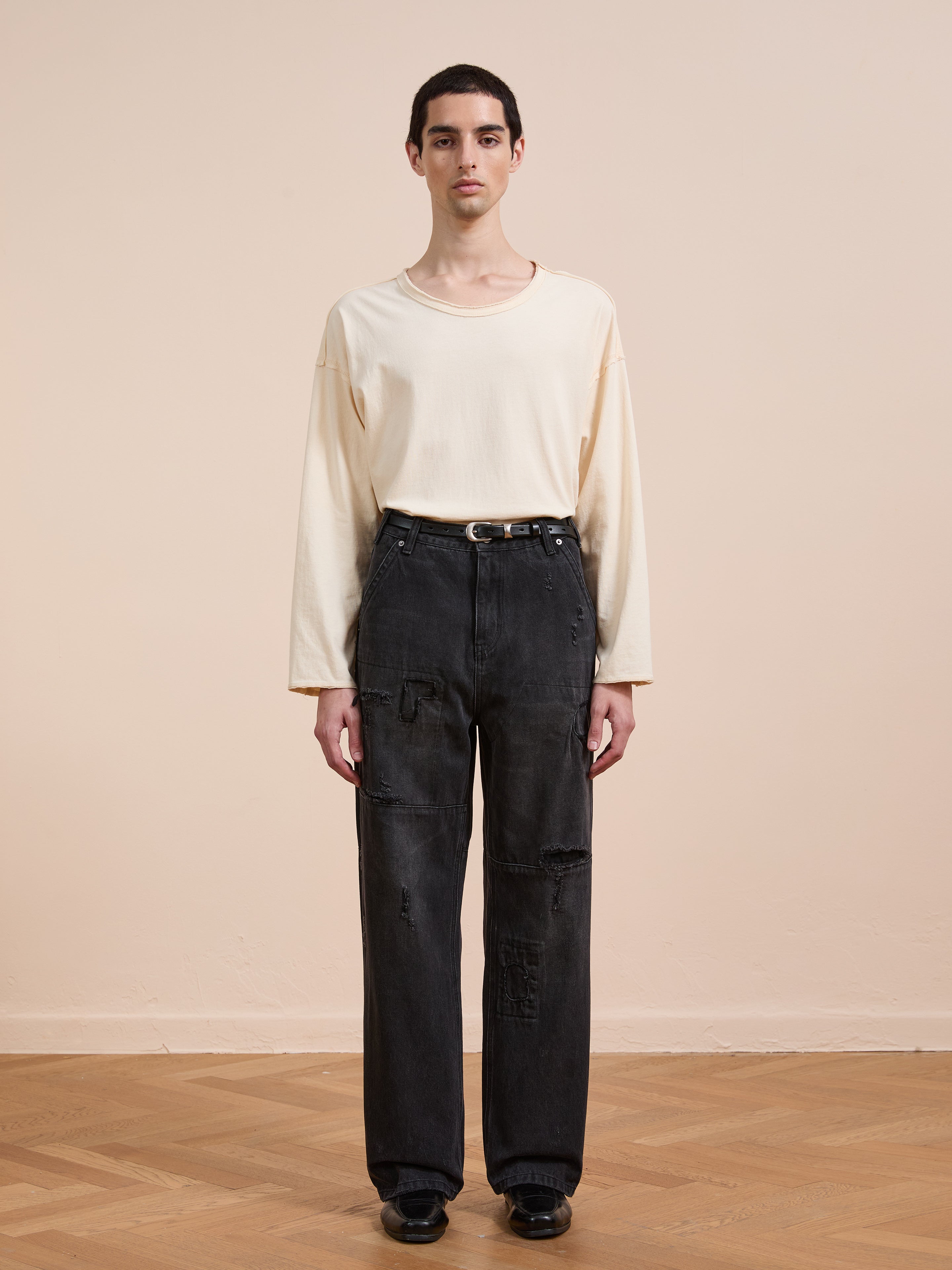 A person stands indoors against a plain beige wall, wearing a loose off-white long-sleeve shirt, FOUND Painter Repaired Jeans with a black belt, and black shoes.