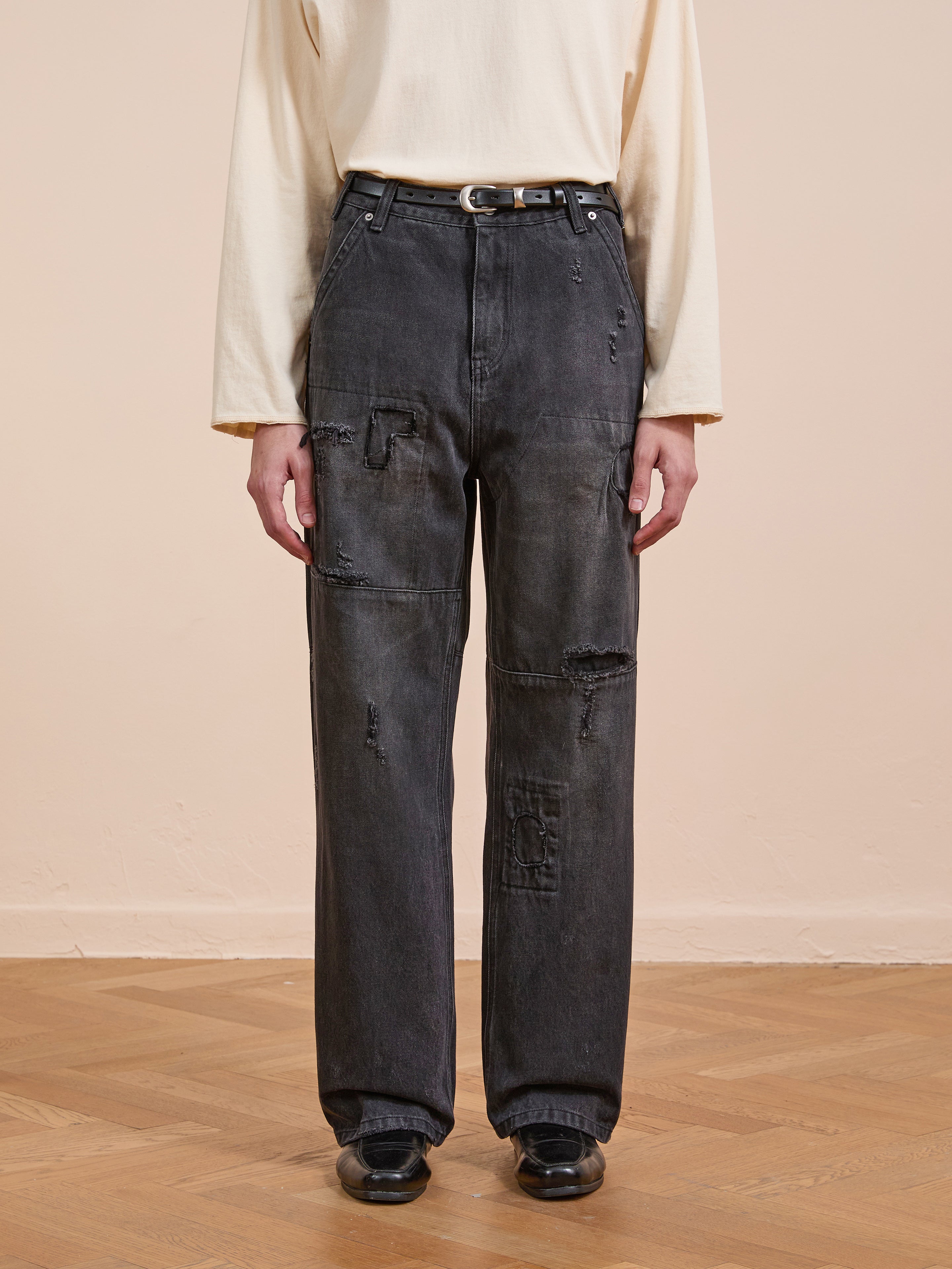 A person stands on a wooden floor against a beige wall, wearing a long-sleeve cream shirt, FOUND Painter Repaired Jeans with a belt, and black shoes.