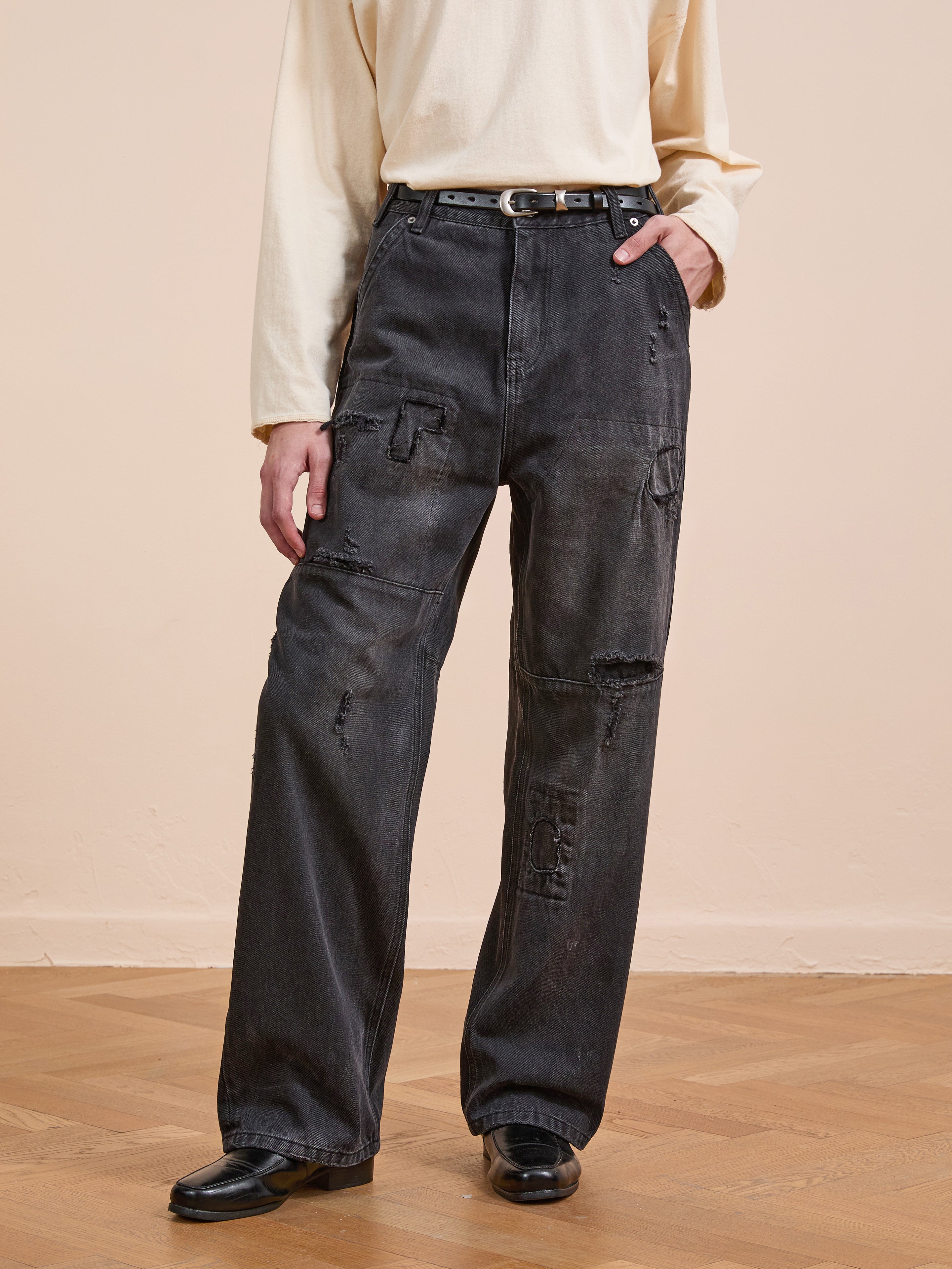 A person wears a cream long-sleeve shirt, FOUND Painter Repaired Jeans with a belt, and black shoes, standing on a wooden floor against a beige wall.