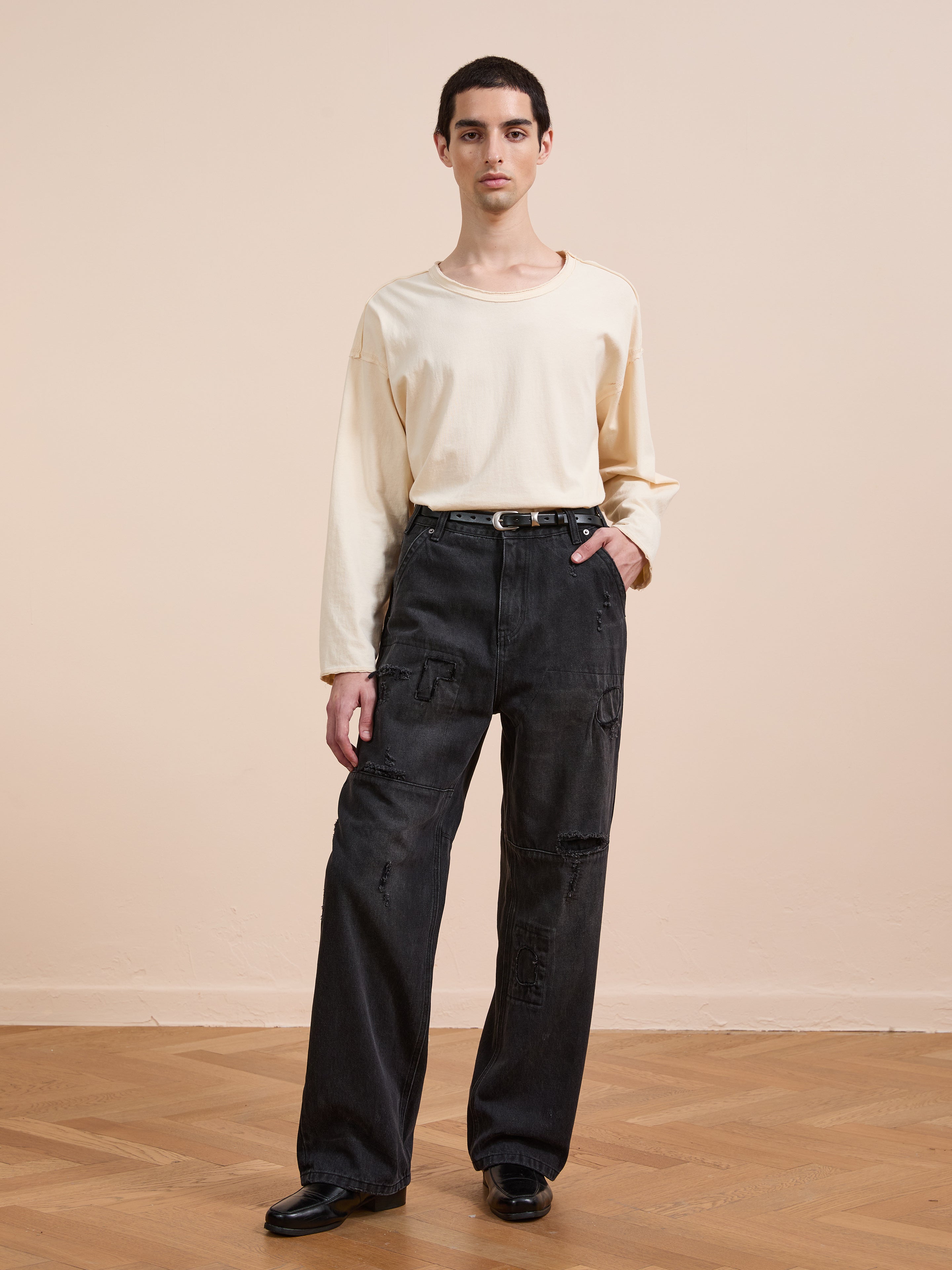 A person stands indoors against a neutral background, wearing a cream long-sleeve shirt, FOUND Painter Repaired Jeans with a black belt, and black shoes.