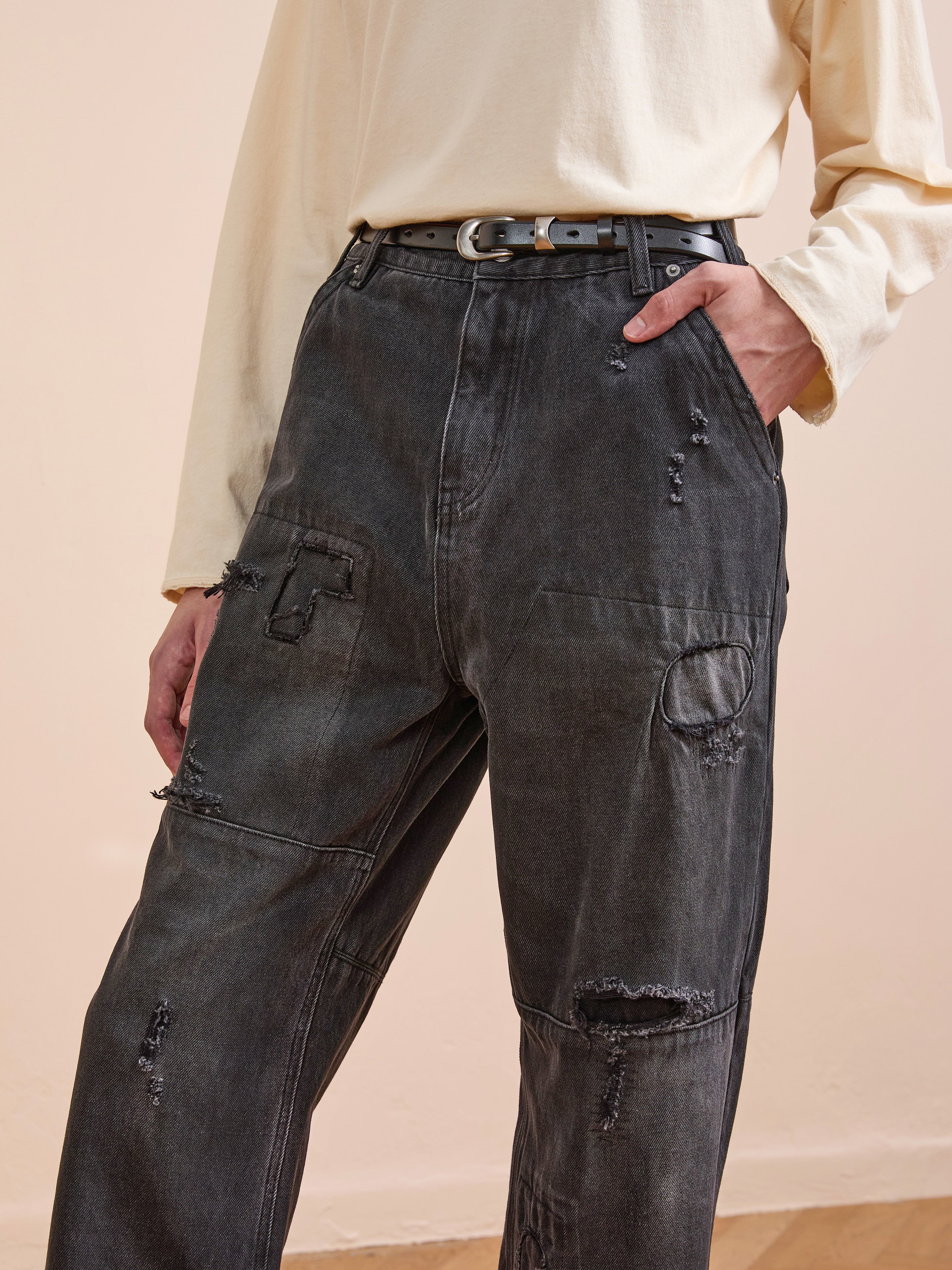 A person wears FOUND Painter Repaired Jeans—a high-waisted black patchwork style—paired with a long-sleeve beige shirt and belt, hand in pocket, for a stylish vintage denim vibe.