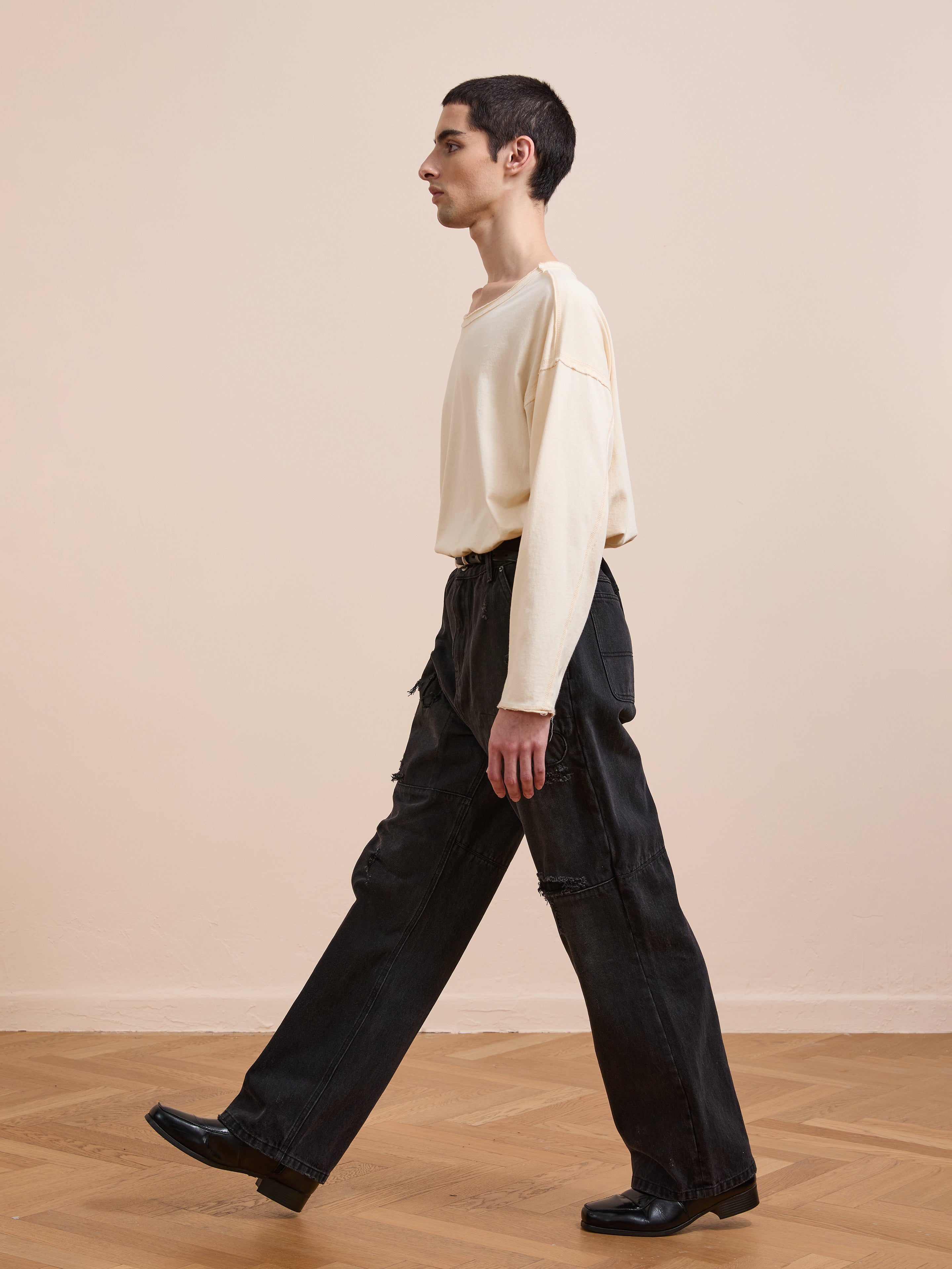 A person with short dark hair walks indoors on a wooden floor, wearing a loose cream long-sleeve shirt, black shoes, and FOUND Painter Repaired Jeans against a plain beige wall.