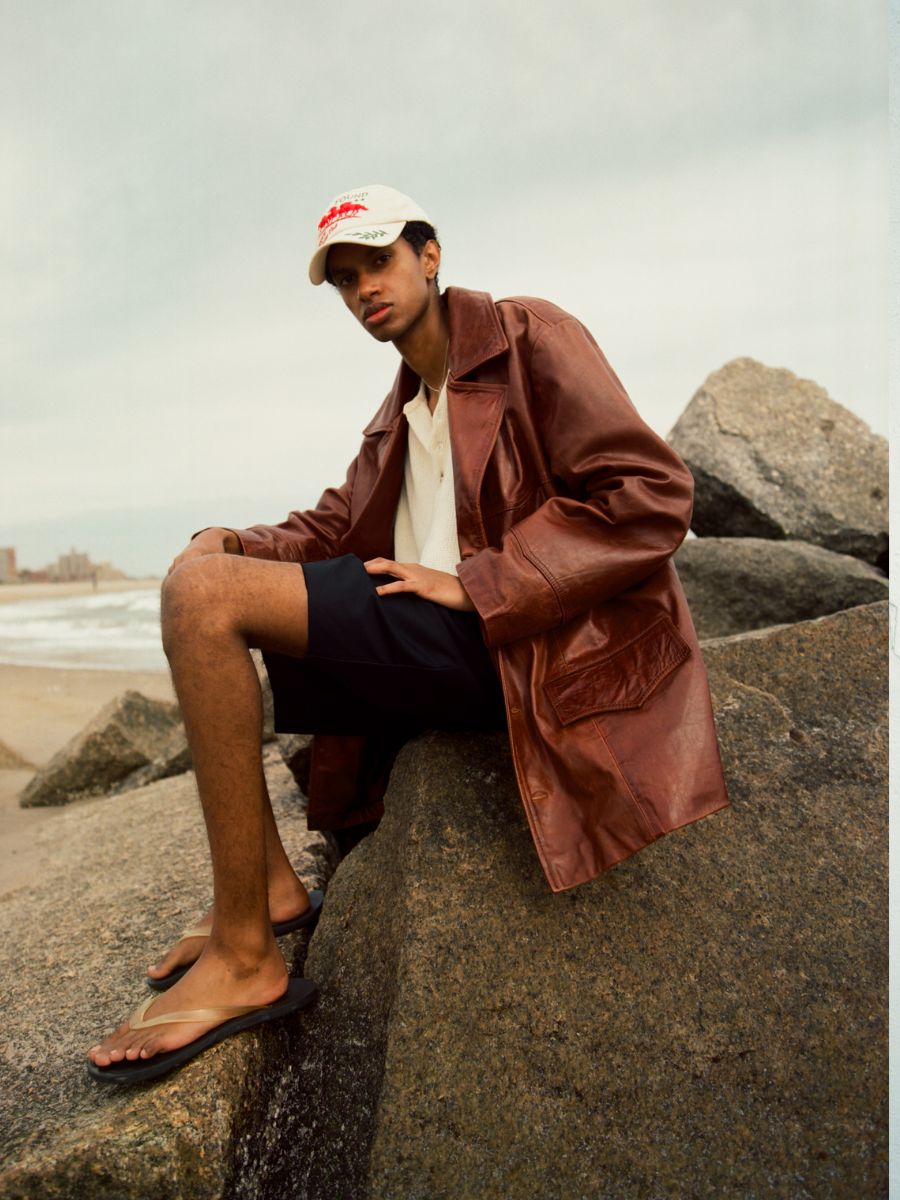 Wearing the FOUND Claret Western Oversized Leather Blazer, a white shirt, dark shorts, sandals, and a white cap, a person sits on large rocks by the beach.
