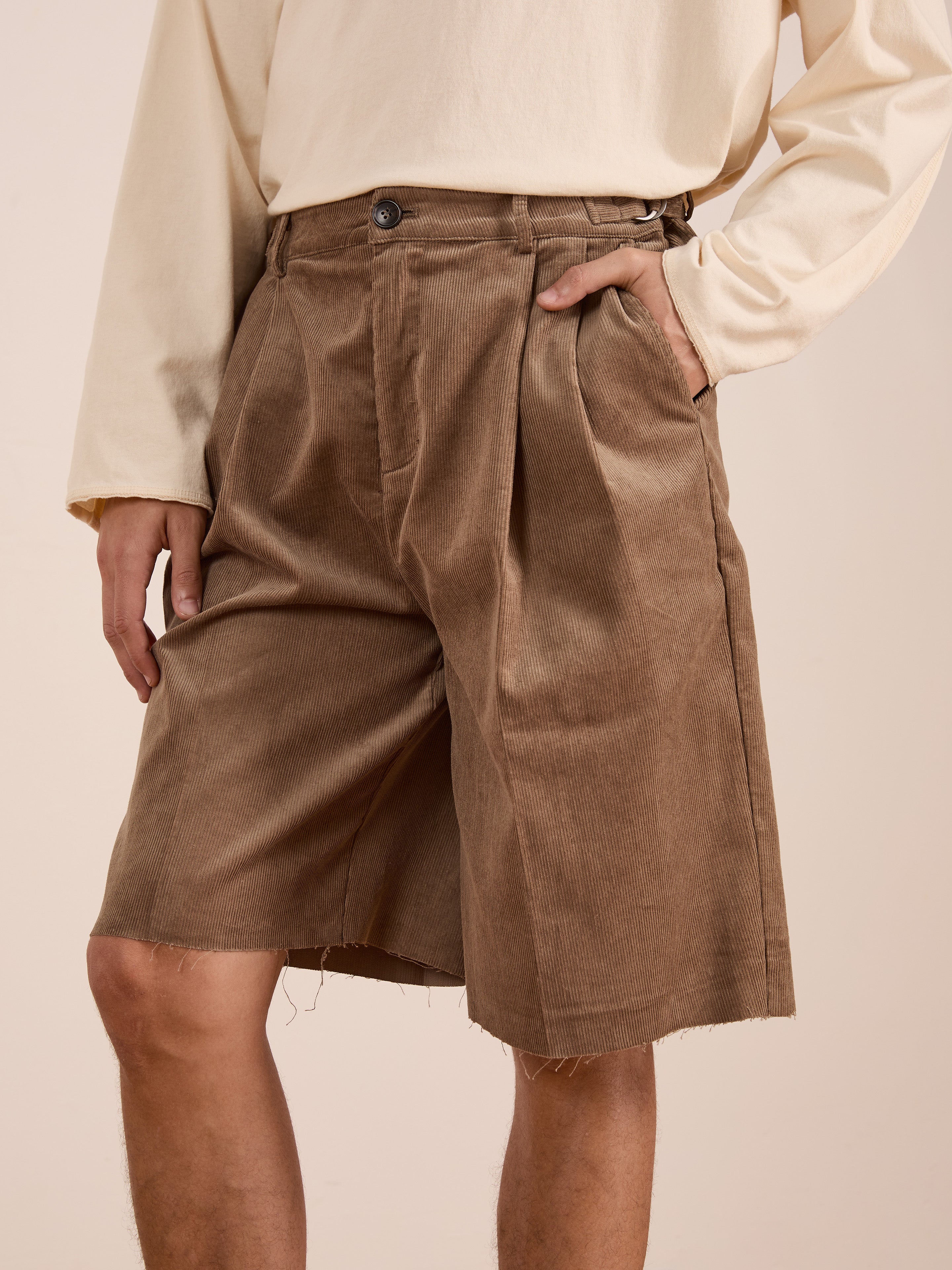 A person wearing a cream long-sleeve shirt and FOUND Corduroy Pleated Trouser Shorts in a relaxed unisex fit with double pleats and raw hems, standing with one hand in their pocket.