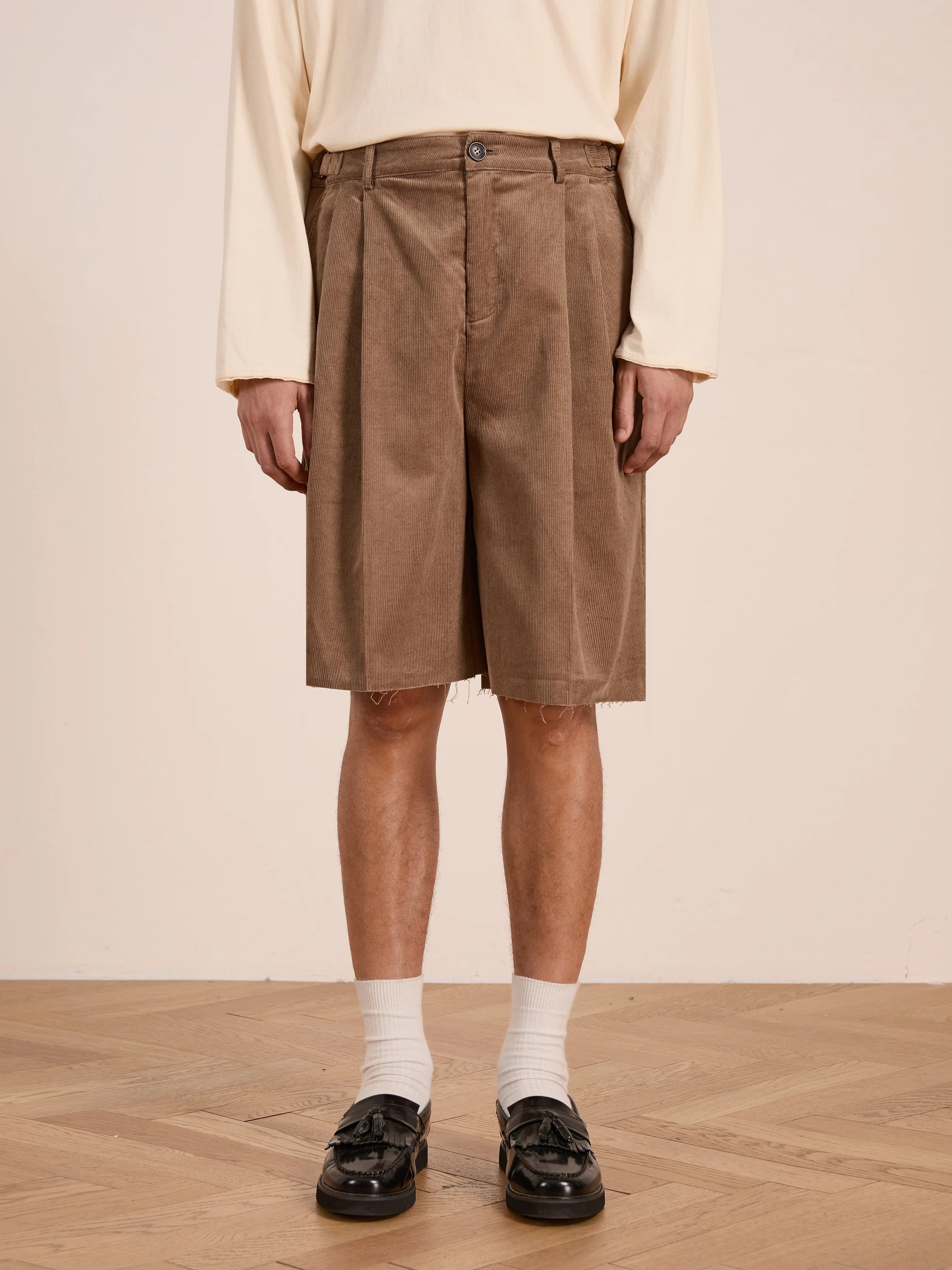 A person wearing a cream long-sleeve shirt, FOUND Corduroy Pleated Trouser Shorts in brown (unisex relaxed fit, double pleated), white socks, and black loafers stands on a wooden floor.
