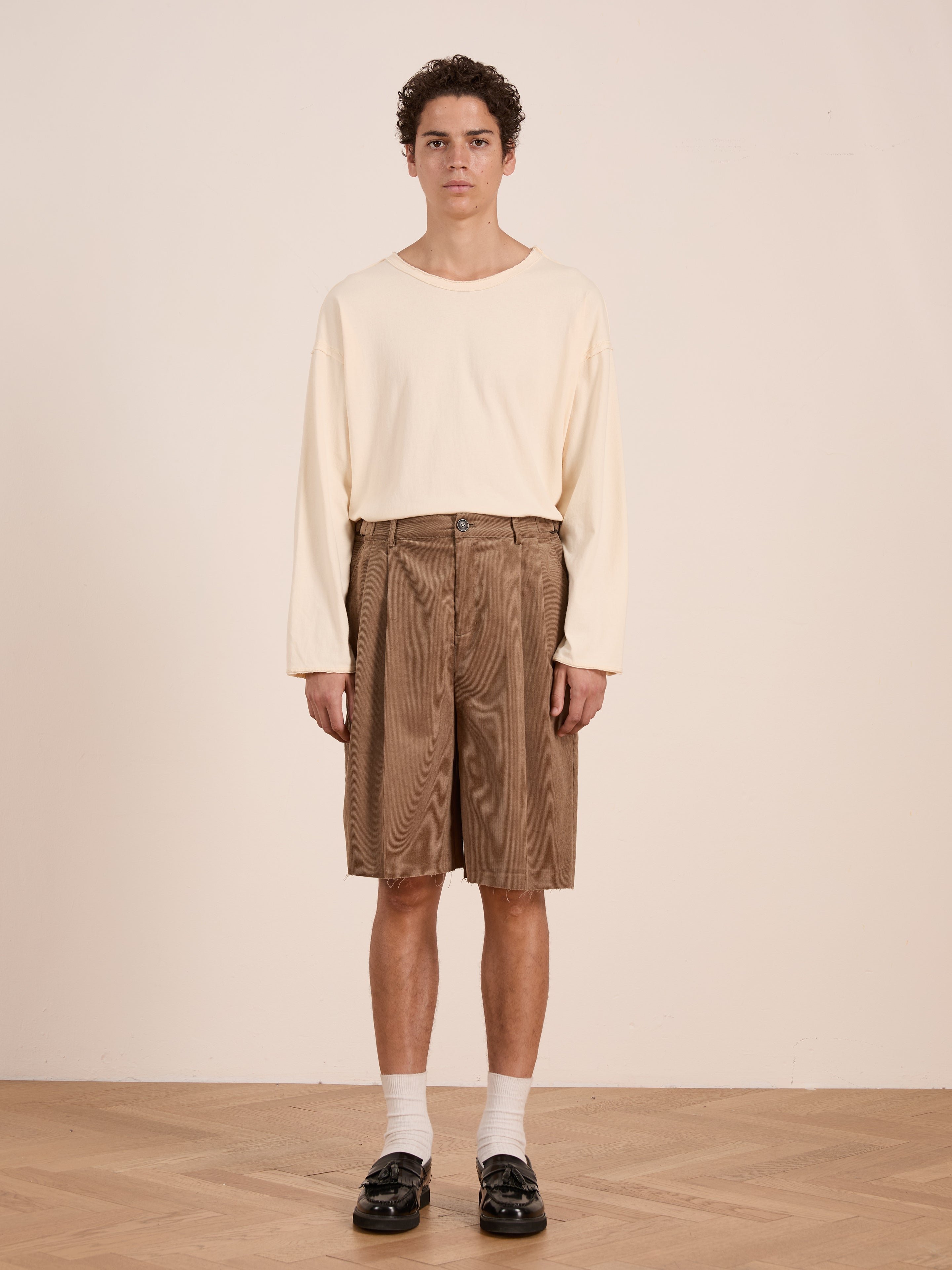 A person stands indoors against a plain background, wearing a cream long-sleeve shirt, FOUND Corduroy Pleated Trouser Shorts in brown (unisex relaxed fit), white socks, and black shoes.