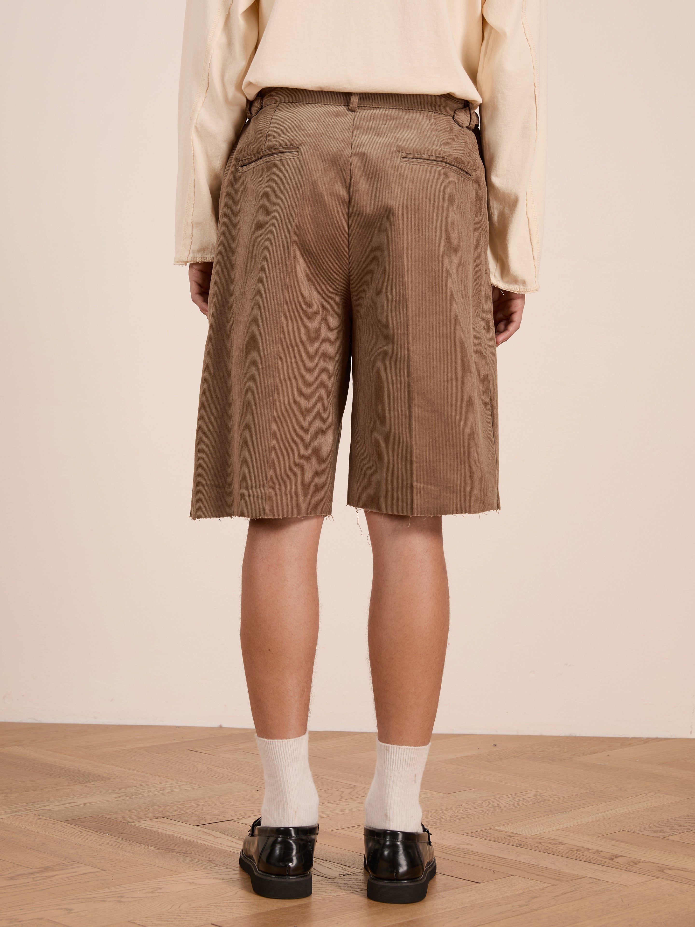 A person wears a beige long-sleeve shirt and FOUND Corduroy Pleated Trouser Shorts, styled with white socks and black shoes, standing on a wooden floor facing away from the camera.