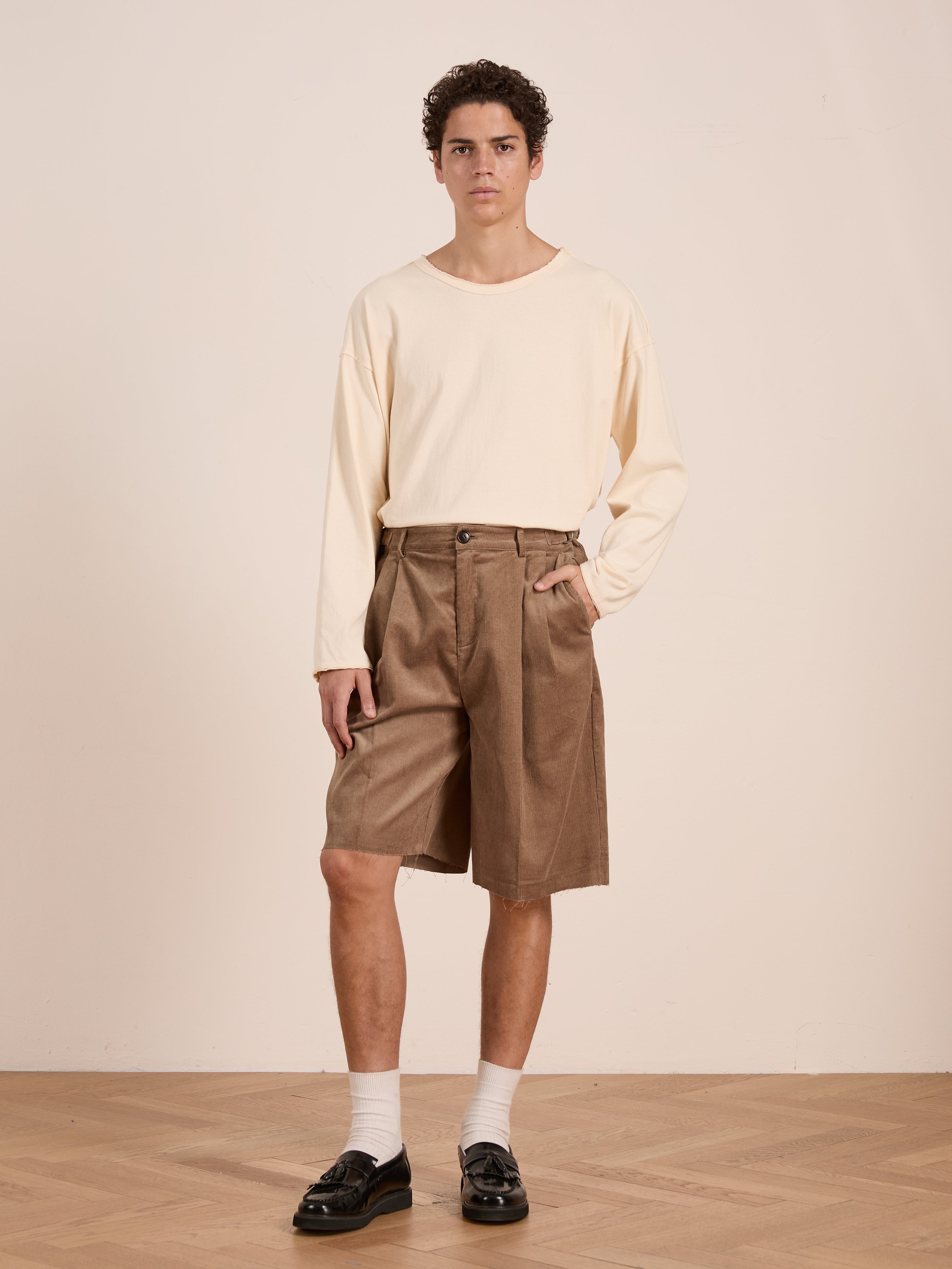 A young person stands indoors in a cream long-sleeve shirt and FOUND Corduroy Pleated Trouser Shorts, featuring a relaxed fit in brown. White socks and black loafers complete the look against a plain, neutral background.