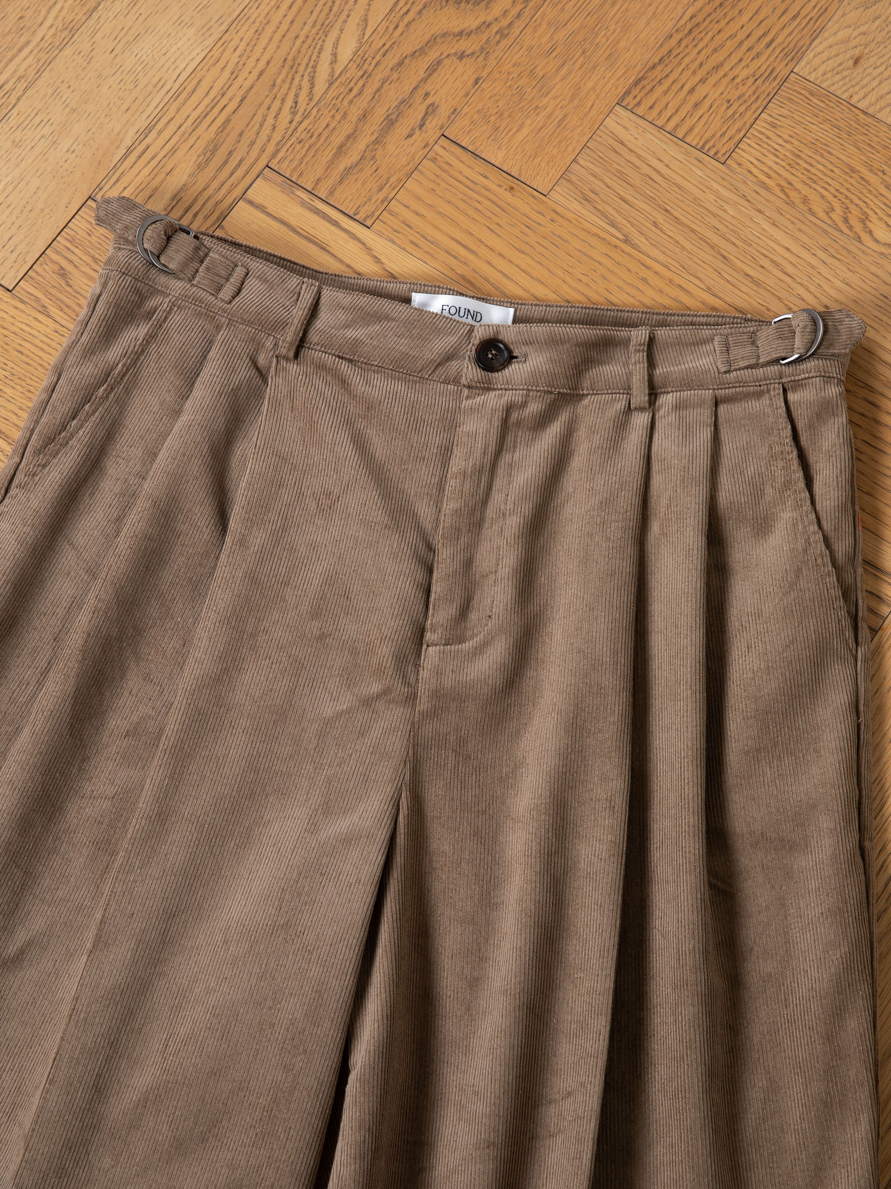 A pair of FOUND Corduroy Pleated Trouser Shorts, featuring a relaxed fit and high waist in brown, is displayed on a wooden herringbone floor.