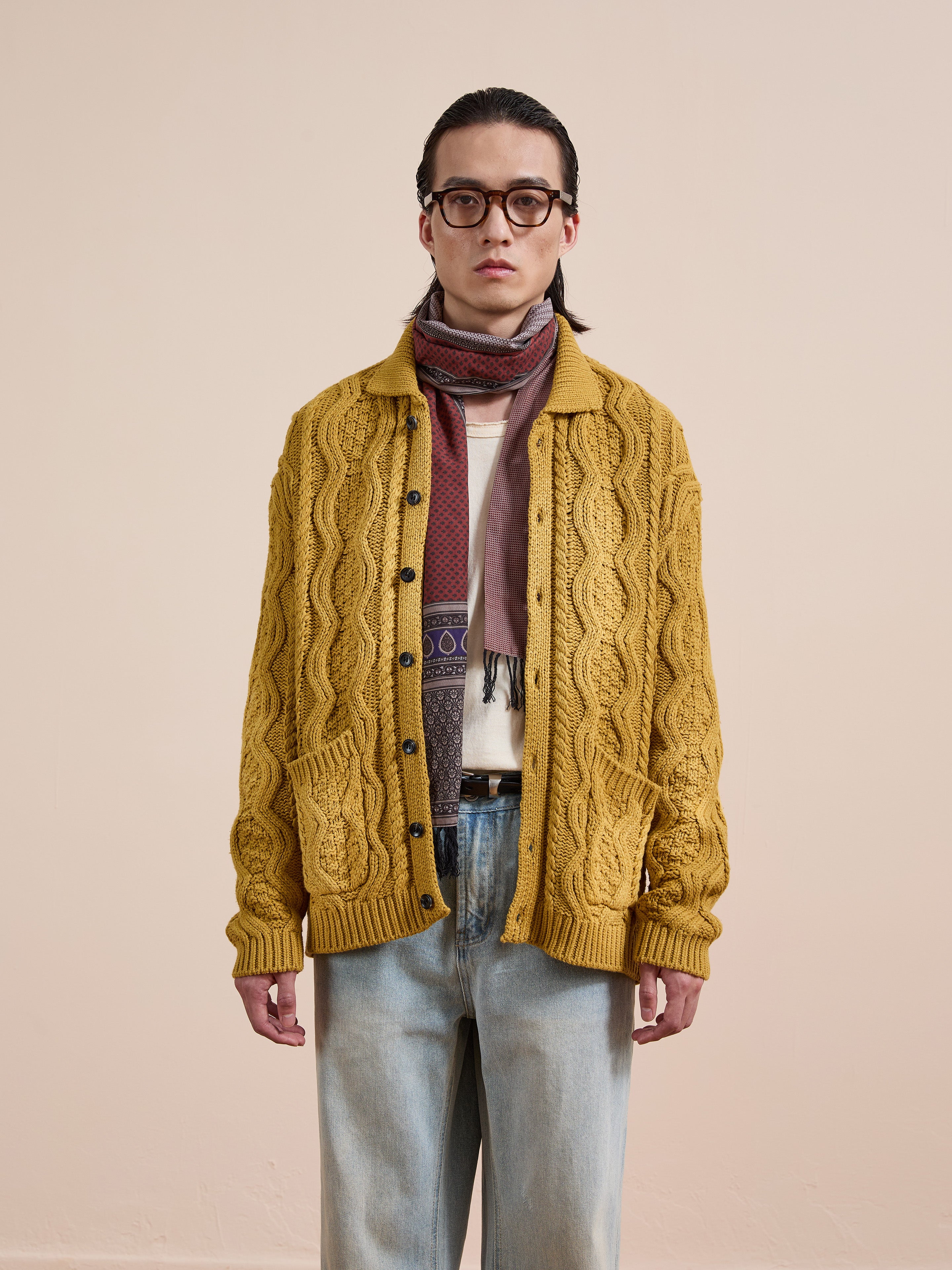 A person wearing glasses, a patterned scarf, a white shirt, light blue jeans, and the FOUND Cable Knit Buttoned Cardigan stands against a plain beige background.