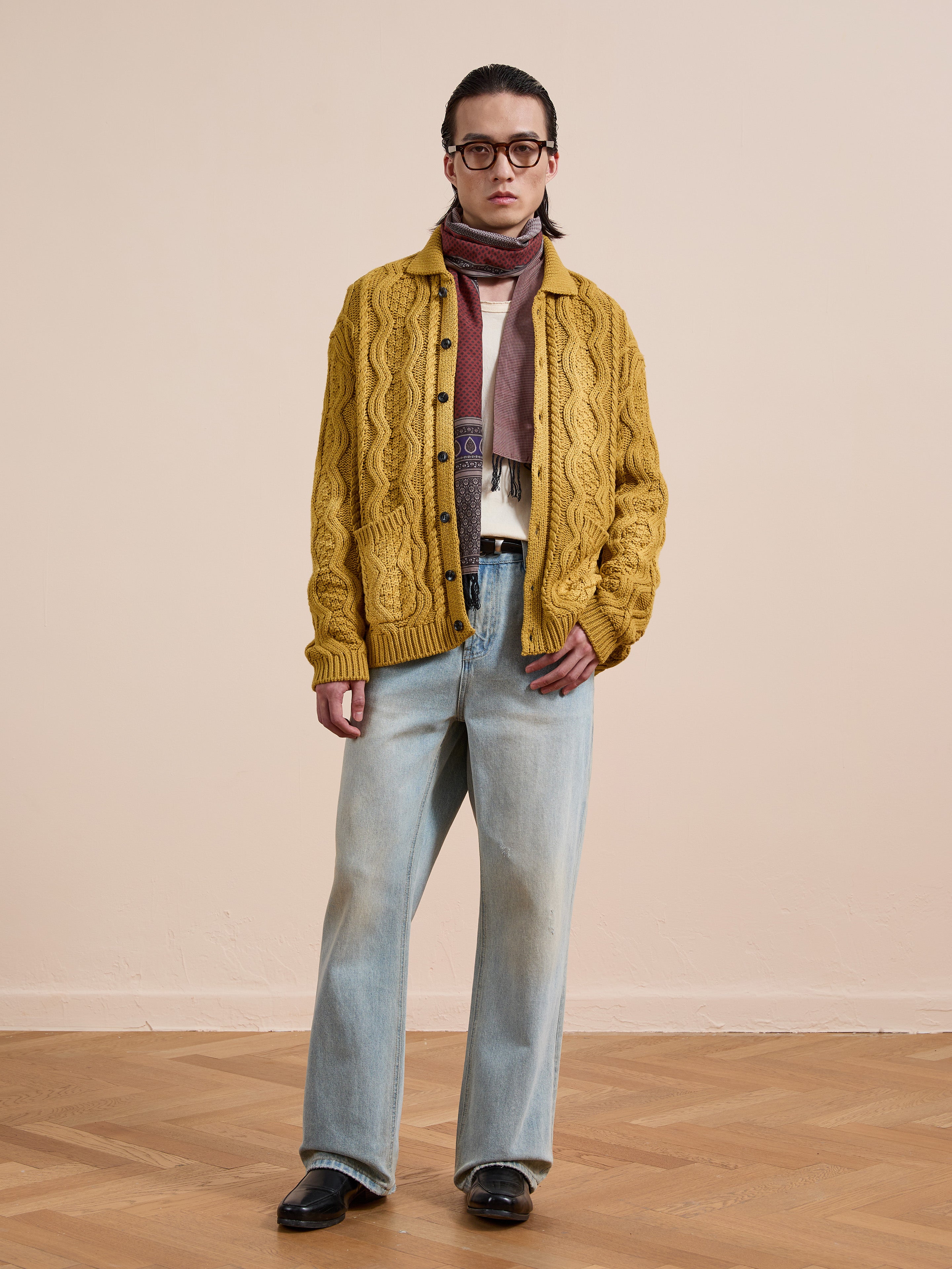 Indoors against a beige wall, a person wears the FOUND Cable Knit Buttoned Cardigan in yellow, paired with light blue jeans, black shoes, glasses, and a multicolored scarf.