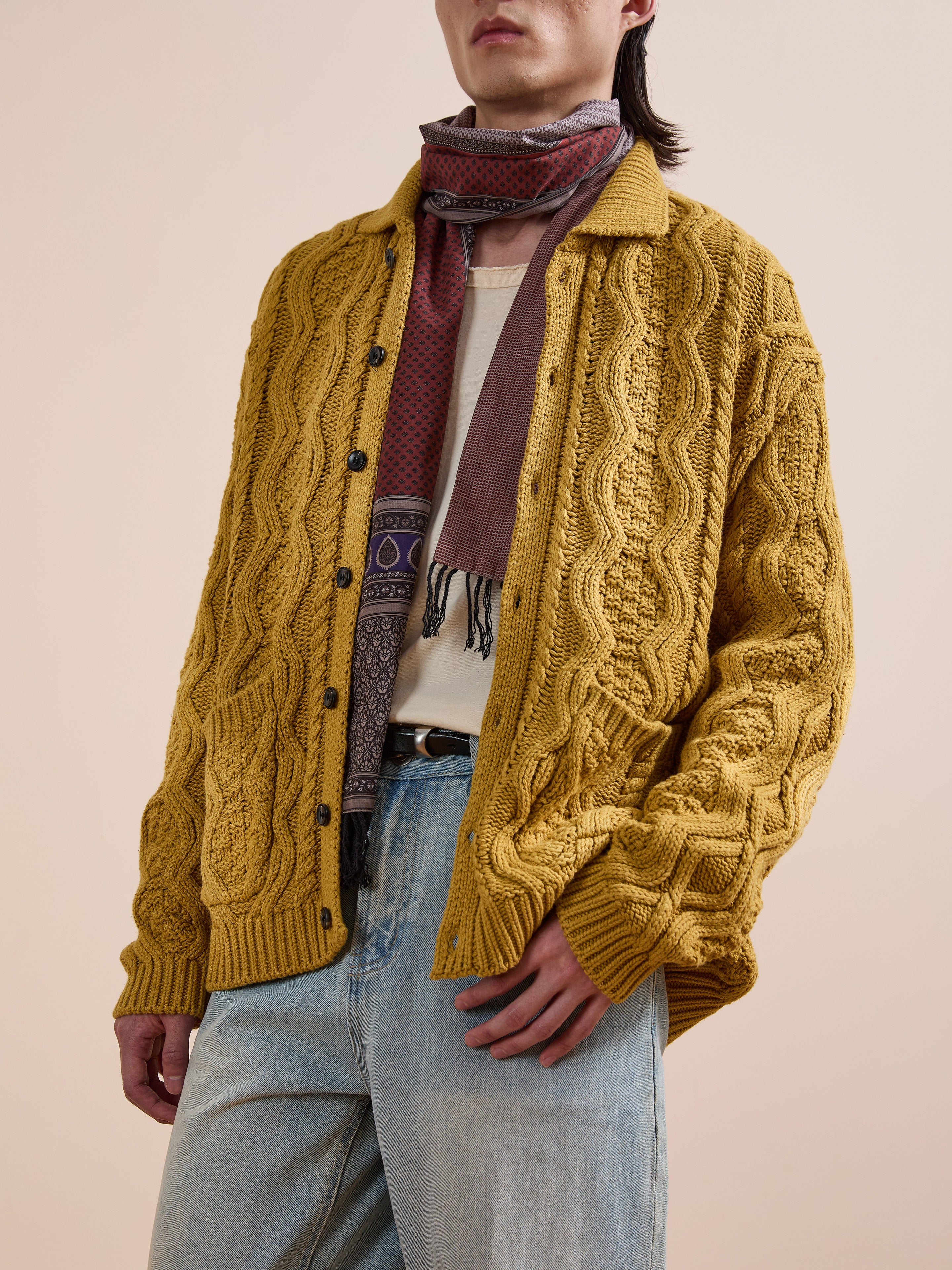 Someone stands against a neutral background wearing the FOUND Cable Knit Buttoned Cardigan in yellow, paired with a patterned scarf, off-white shirt, and light blue jeans.