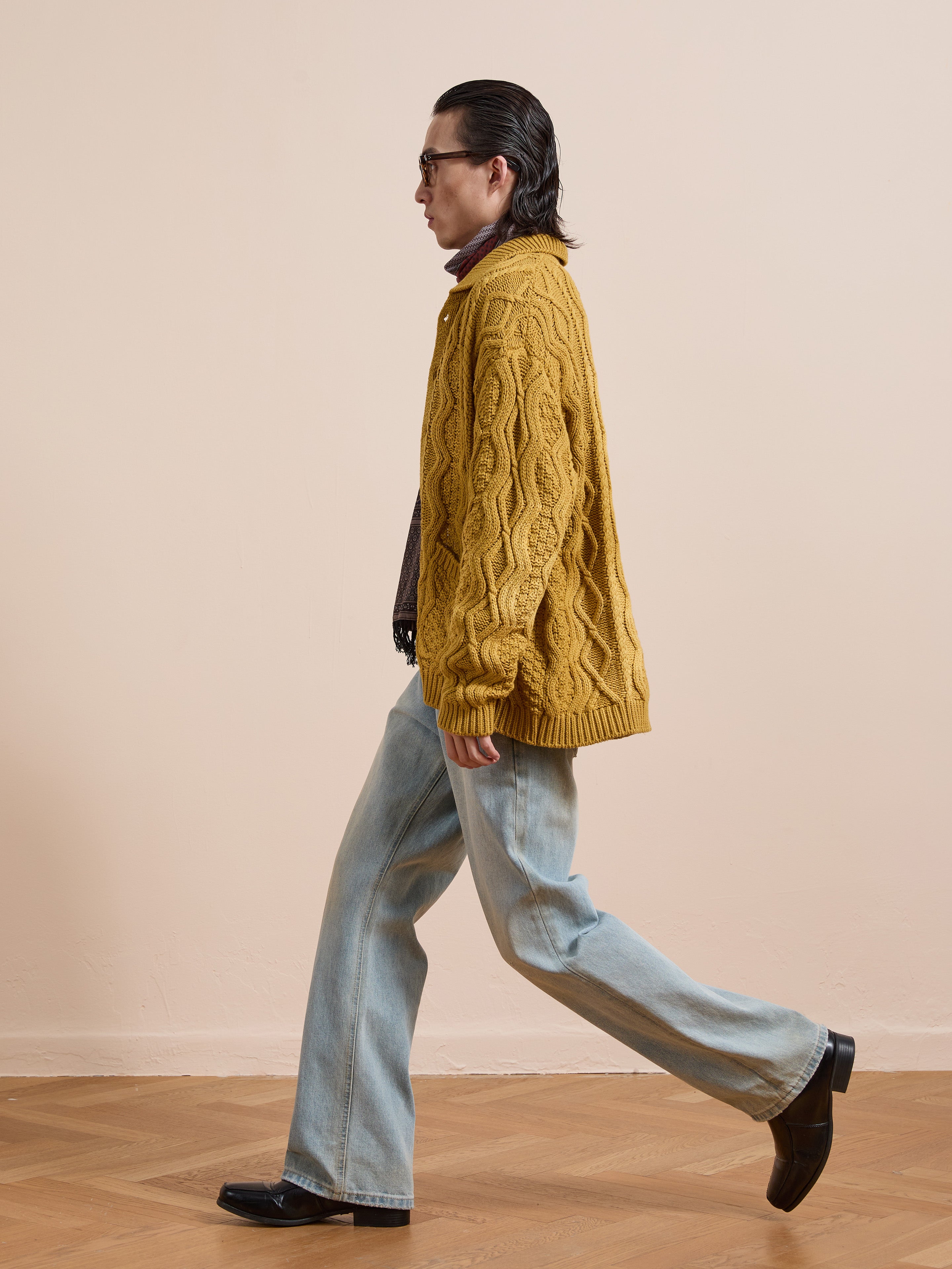 Someone walks indoors on a wooden floor against a beige wall, wearing the FOUND Cable Knit Buttoned Cardigan in mustard yellow with chunky cables, paired with light blue wide-leg jeans and black shoes.