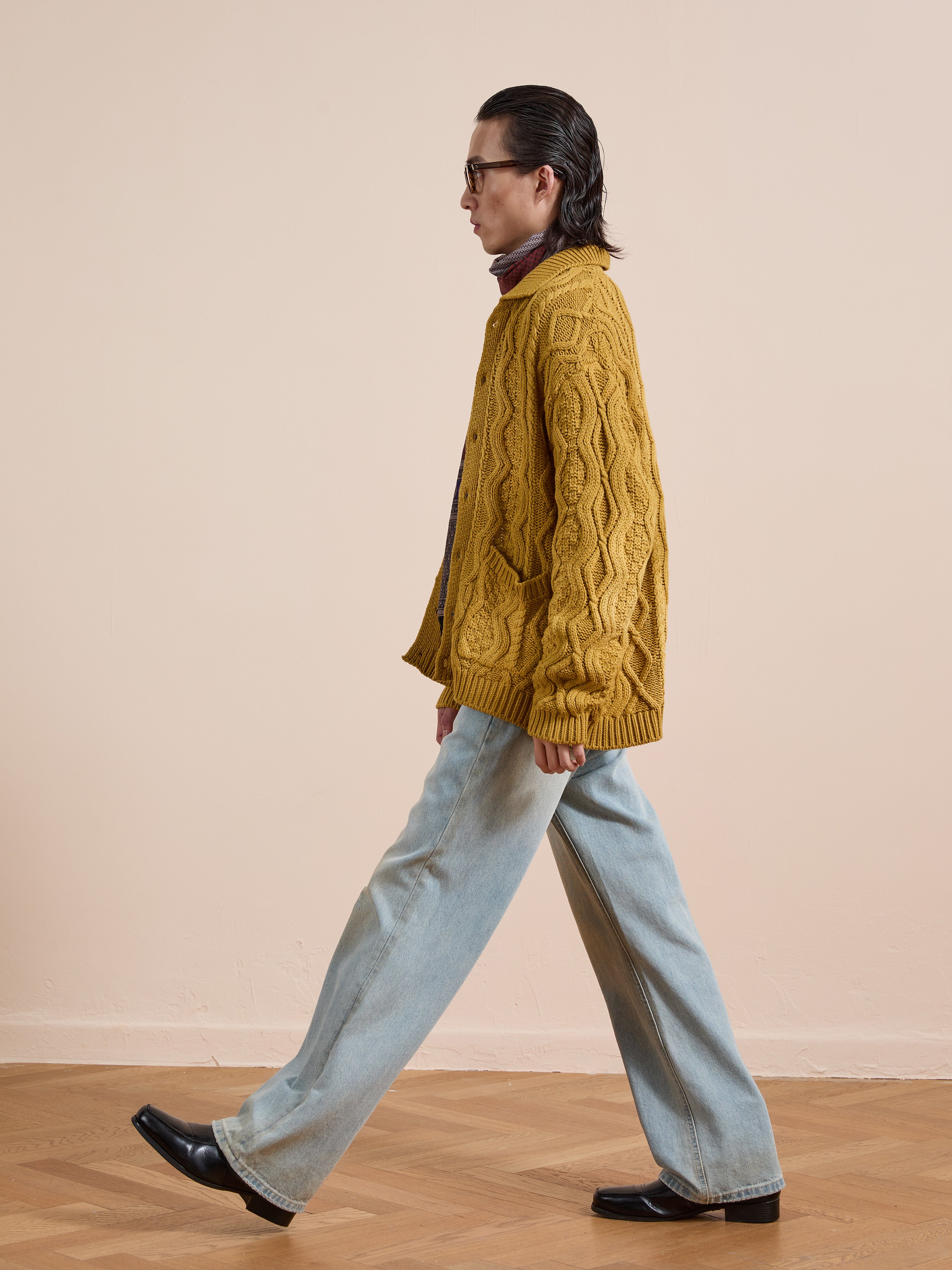 A person with long dark hair walks sideways, wearing the FOUND Cable Knit Buttoned Cardigan in mustard yellow, light blue wide-leg jeans, black shoes, and glasses on a wooden floor against a beige wall.