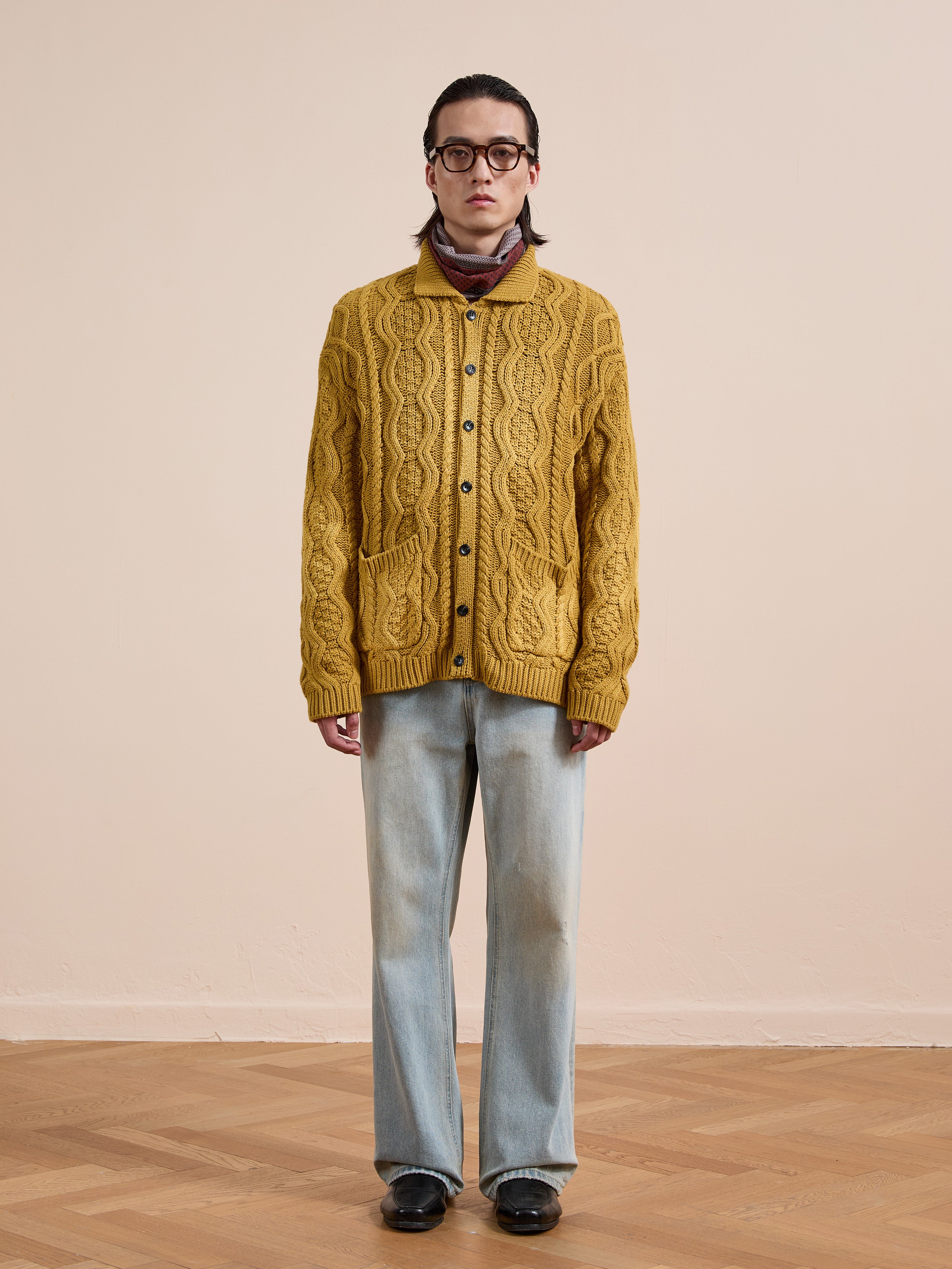 Indoors against a neutral background, a person wears the FOUND Cable Knit Buttoned Cardigan in mustard yellow with chunky cables, paired with light blue jeans, black shoes, glasses, and a neck scarf.
