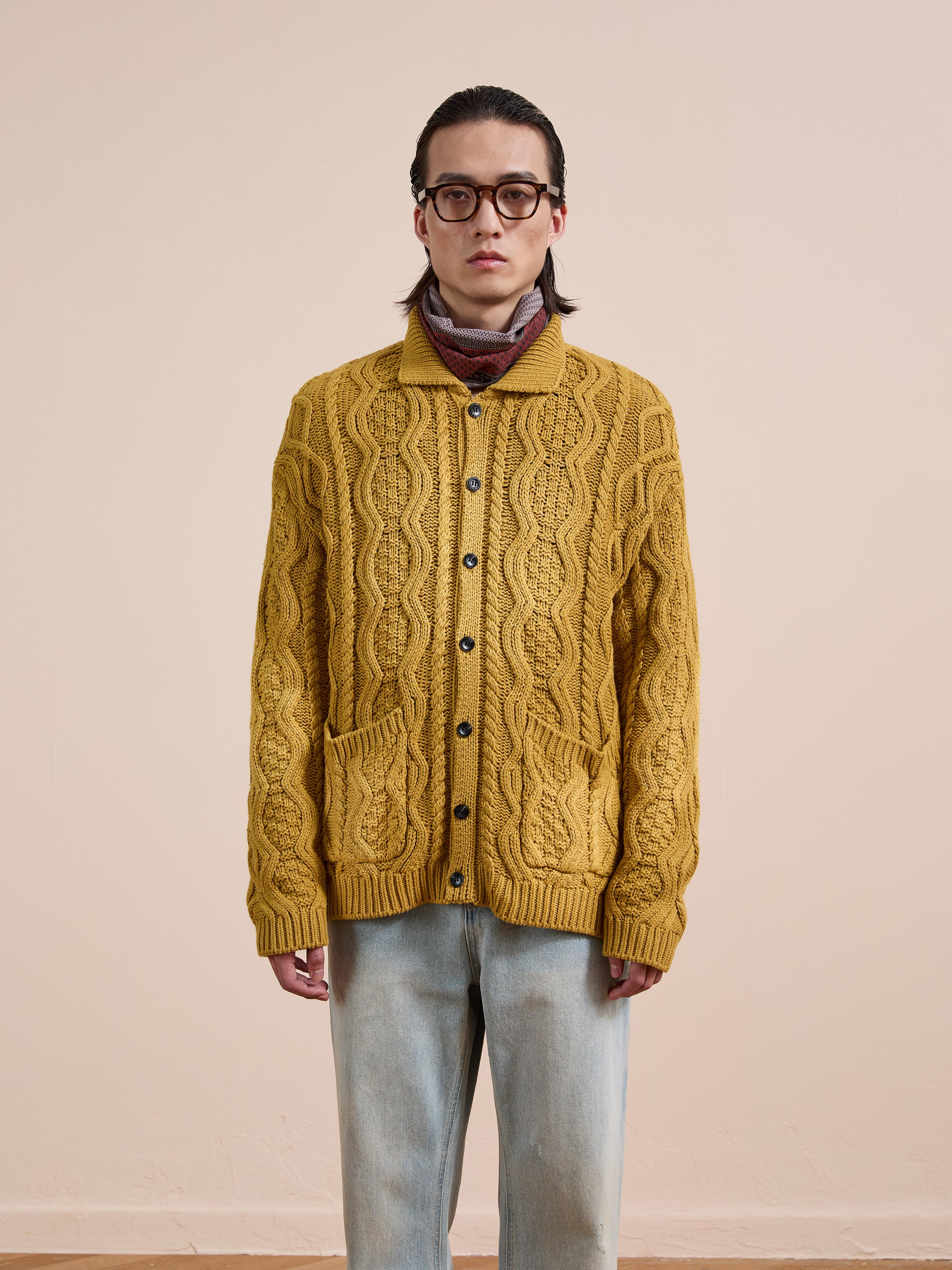 A person wearing glasses, a scarf, and light blue jeans models the FOUND Cable Knit Buttoned Cardigan in mustard yellow with front pockets, standing against a plain beige background.