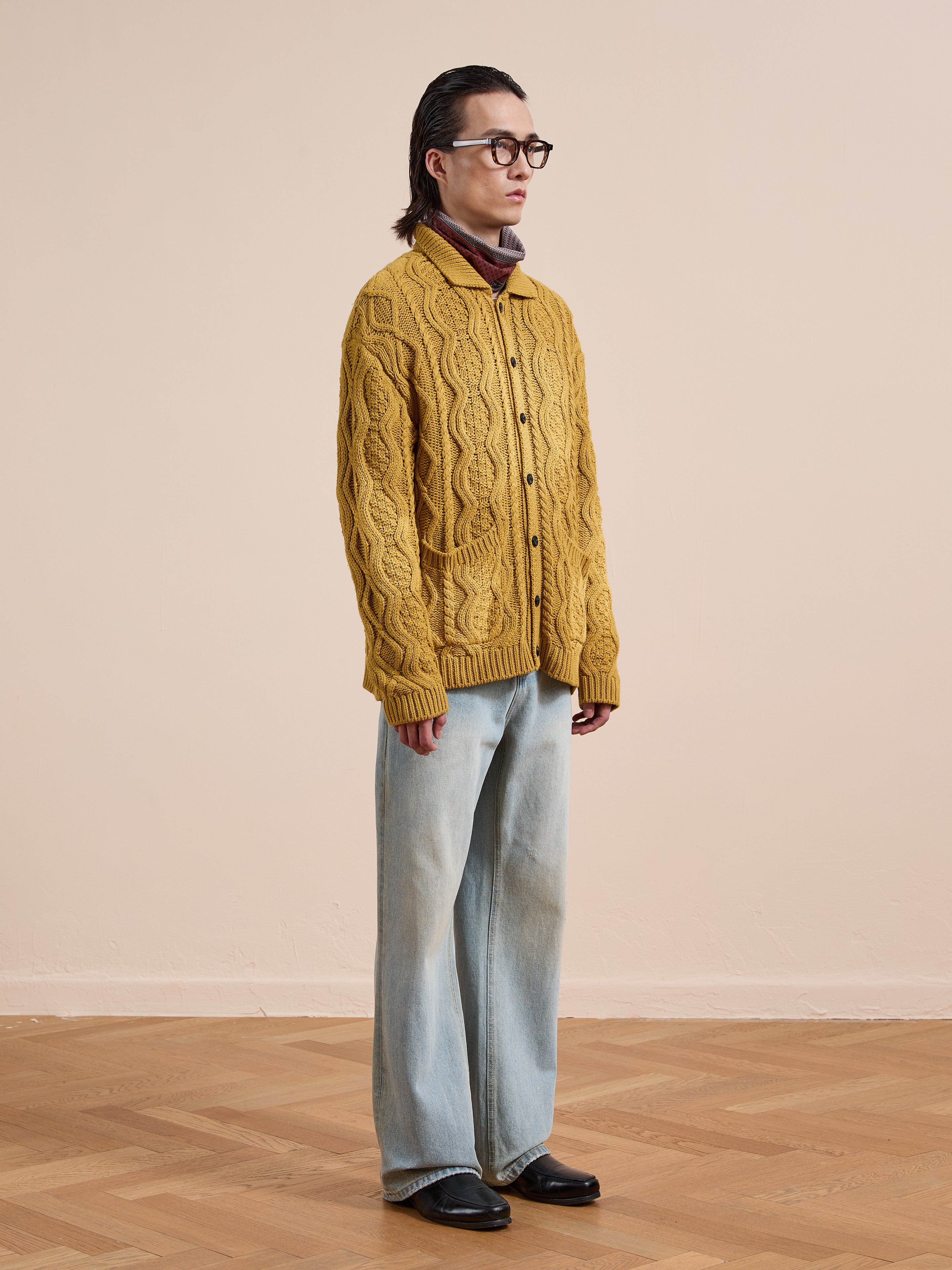 Person stands indoors against a beige background, wearing the FOUND Cable Knit Buttoned Cardigan in yellow with chunky cable knitting, paired with light blue jeans, black shoes, glasses, and a maroon turtleneck.