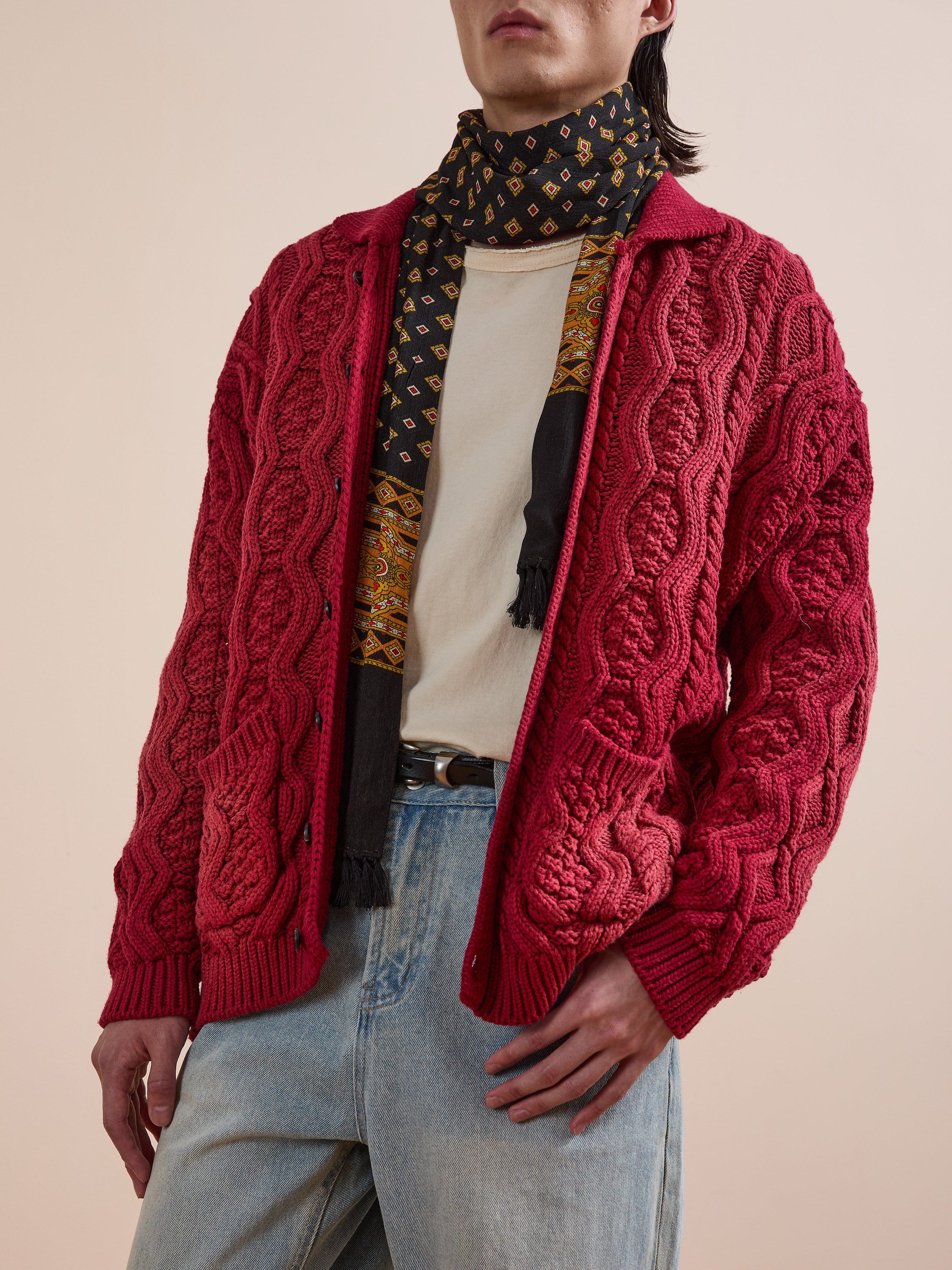 A person in a FOUND Cable Knit Buttoned Cardigan, printed scarf, white tee, and light blue jeans stands with one hand in their pocket against a plain background.