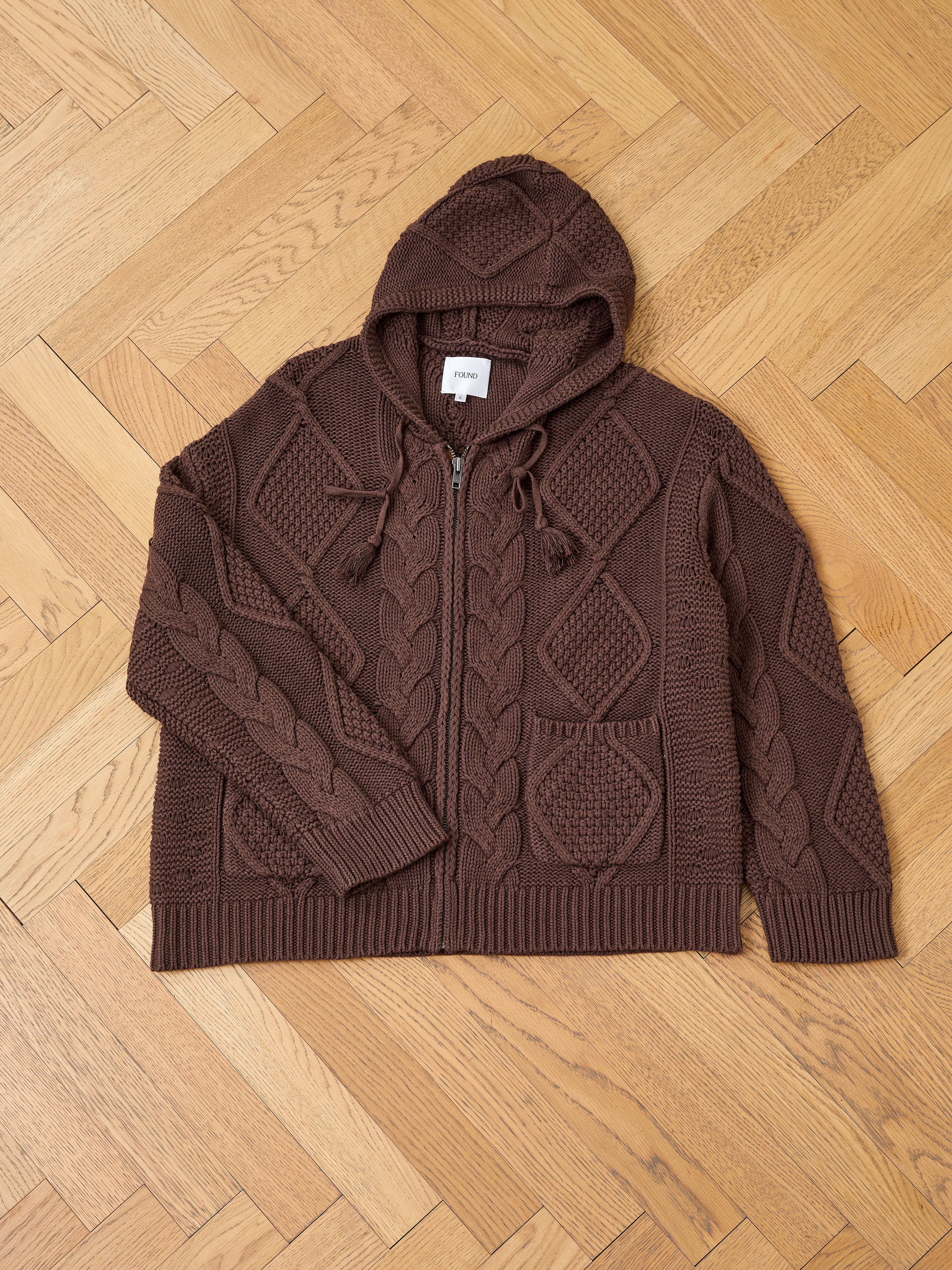 The FOUND Zip Up Cable Knit Hoodie, a unisex brown hoodie with front pockets and vintage-inspired cable knit details, is shown laid flat on a light wood herringbone floor, offering a relaxed fit and timeless style.