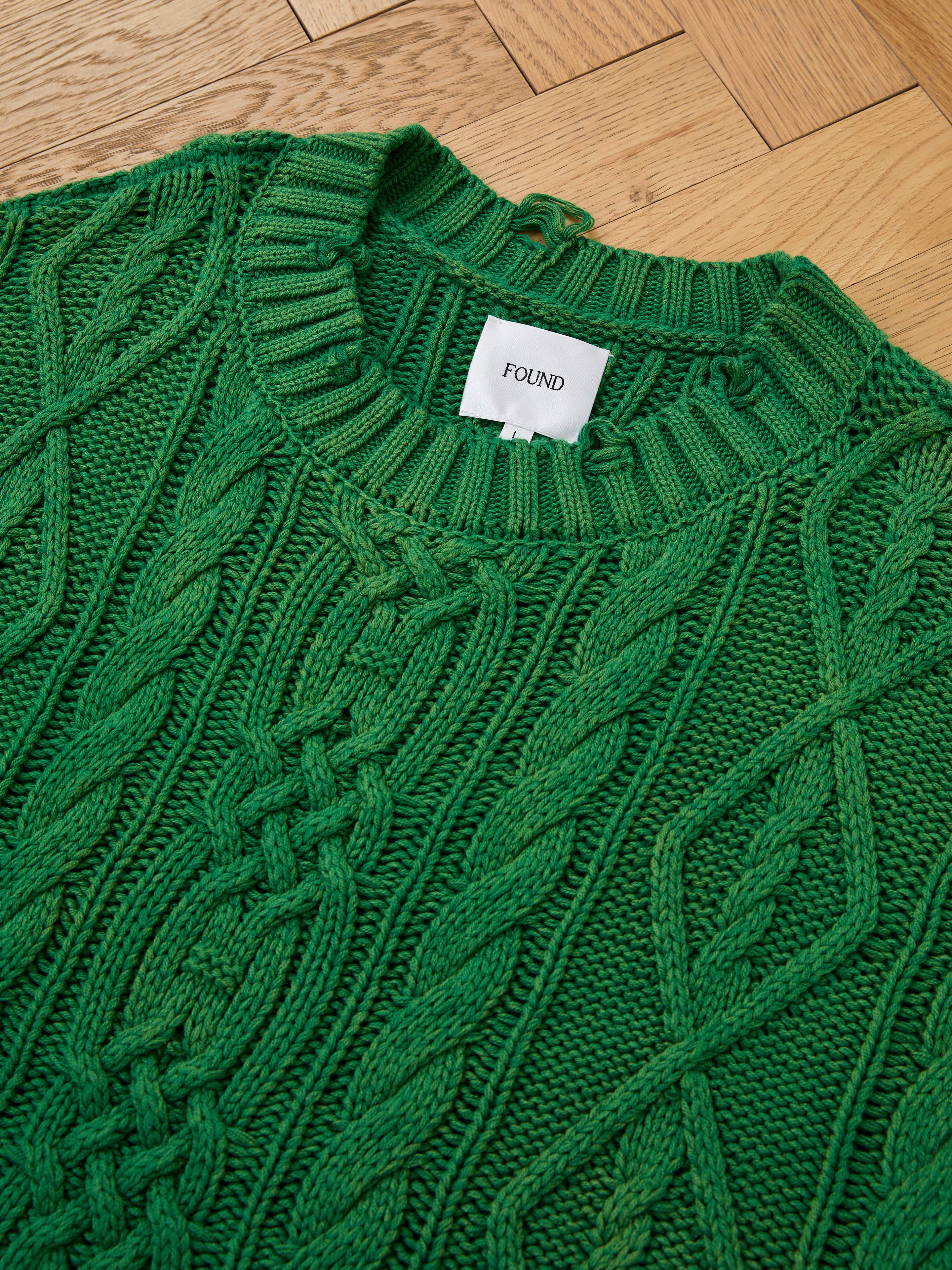 The Cable Knit Pullover Sweater by Found is a green unisex knit, shown flat on a wooden floor, offering cozy comfort and versatile style.