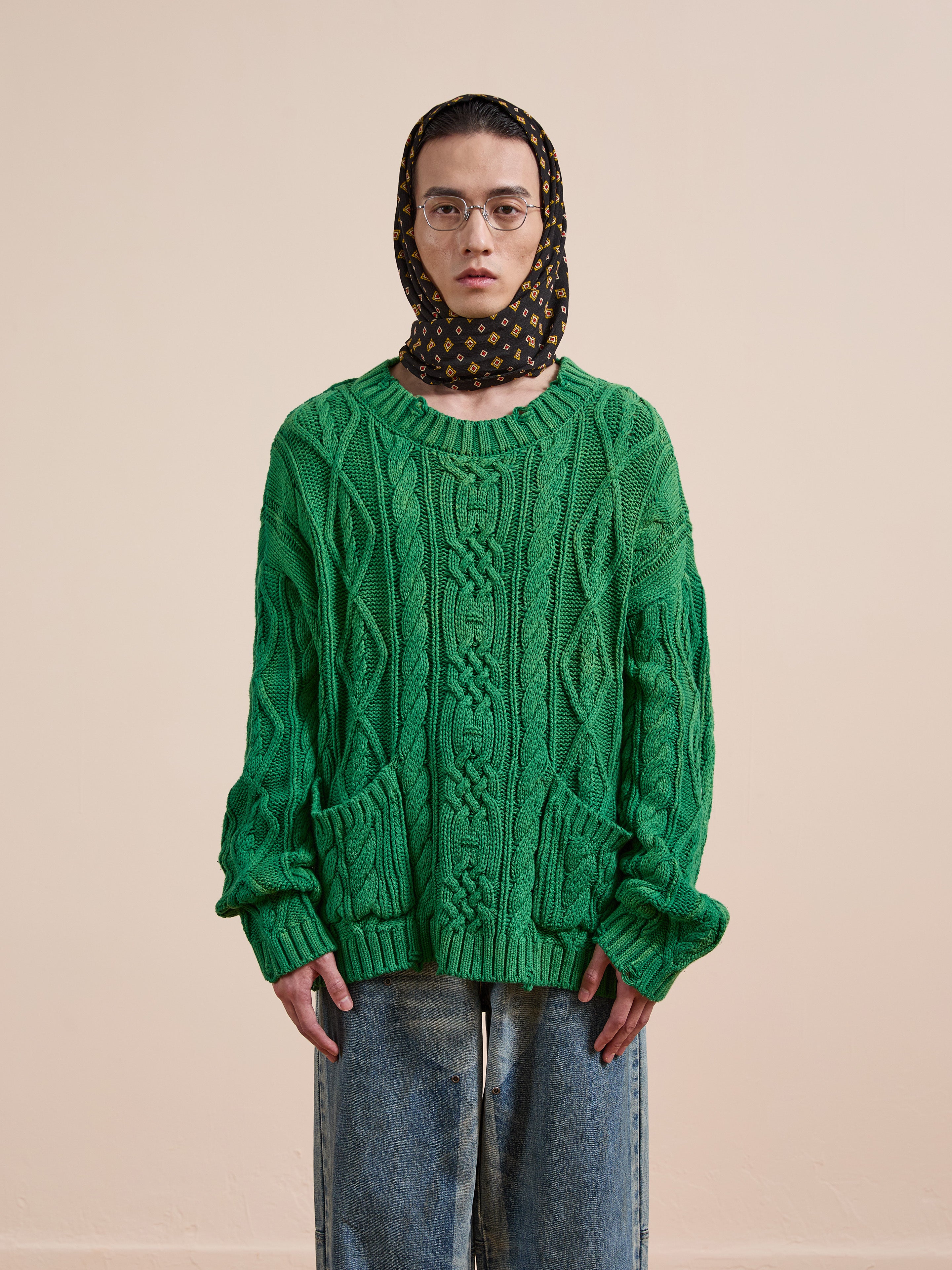 A person in Found's green Cable Knit Pullover Sweater, loose jeans, glasses, and a patterned headscarf stands against a plain beige background.