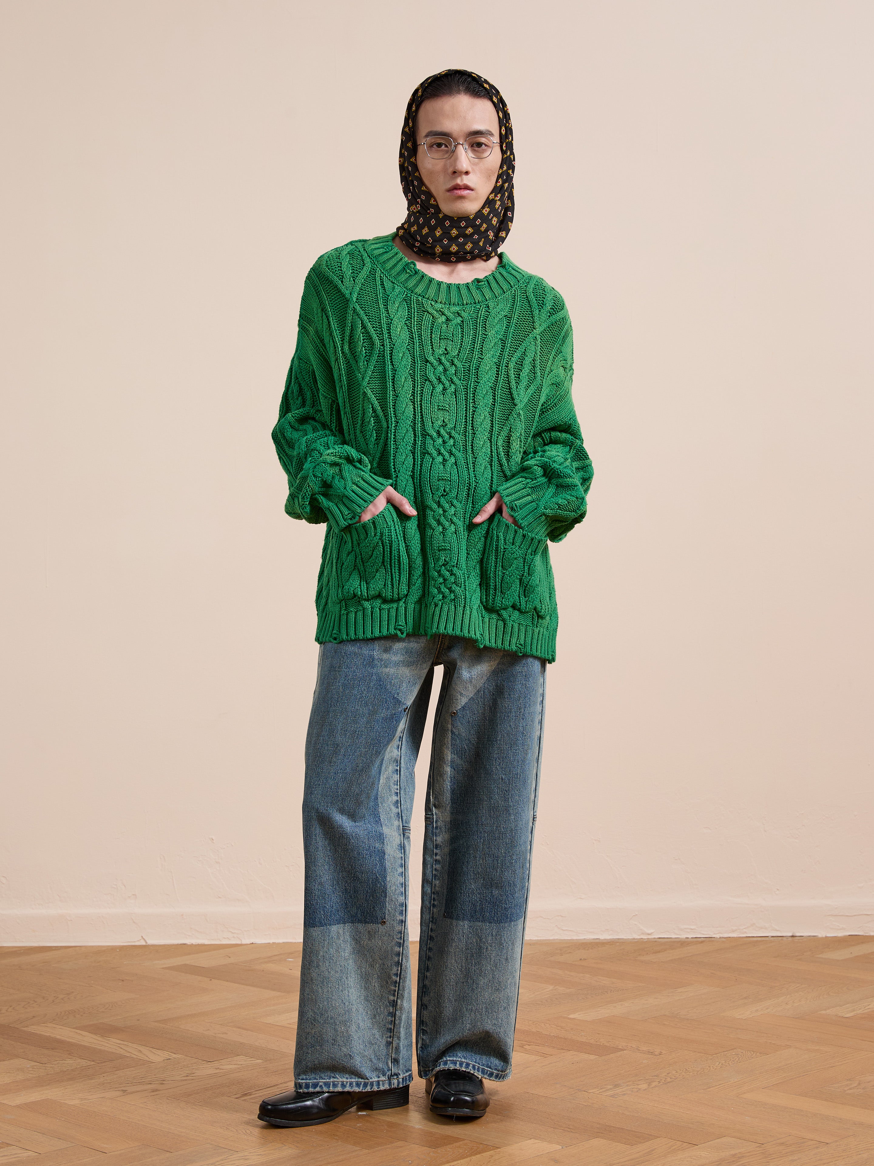 A person stands indoors wearing Found's Cable Knit Pullover Sweater in green, wide-leg jeans, black shoes, and a black polka dot headscarf, with hands tucked into the sweater pockets.