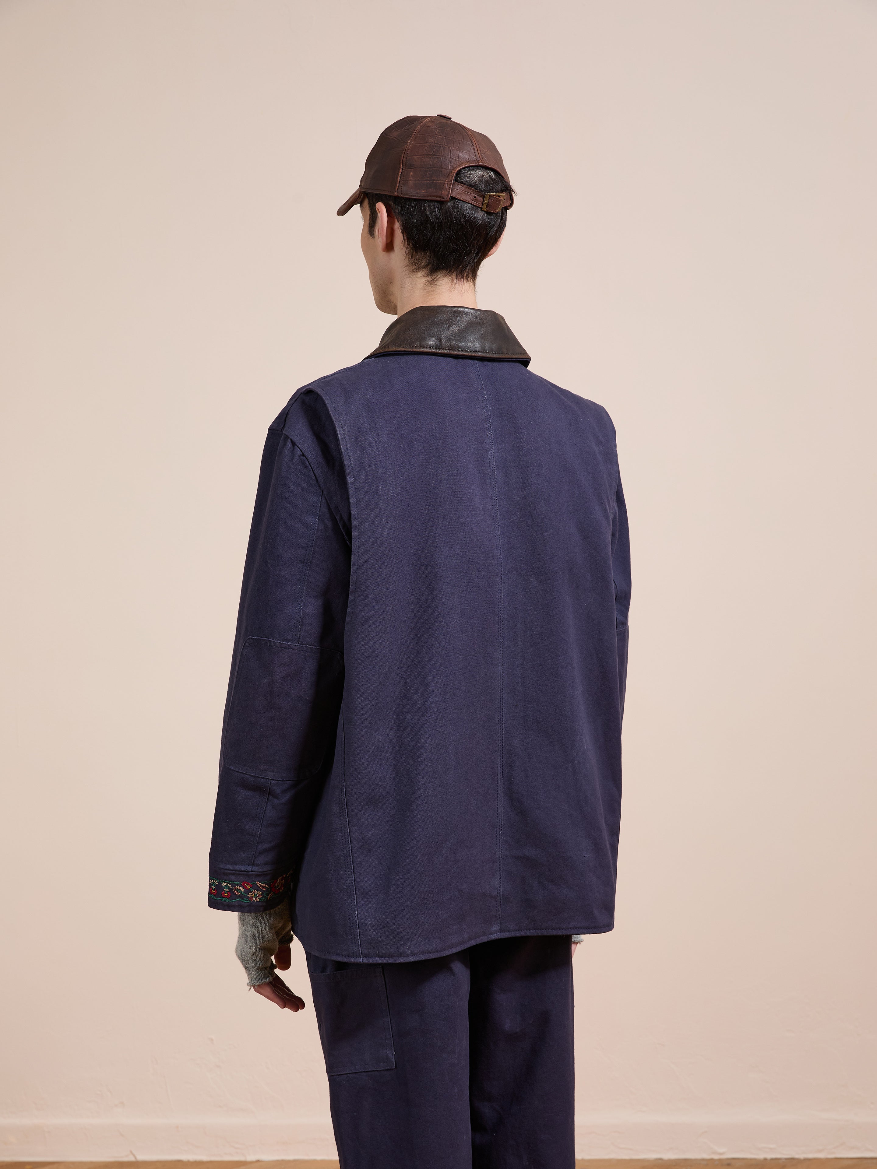 A person wears the Found Canvas Distressed Barn Jacket with a leather collar, matching pants, and a brown cap, styled in a unisex relaxed fit, photographed from behind against a plain light background.