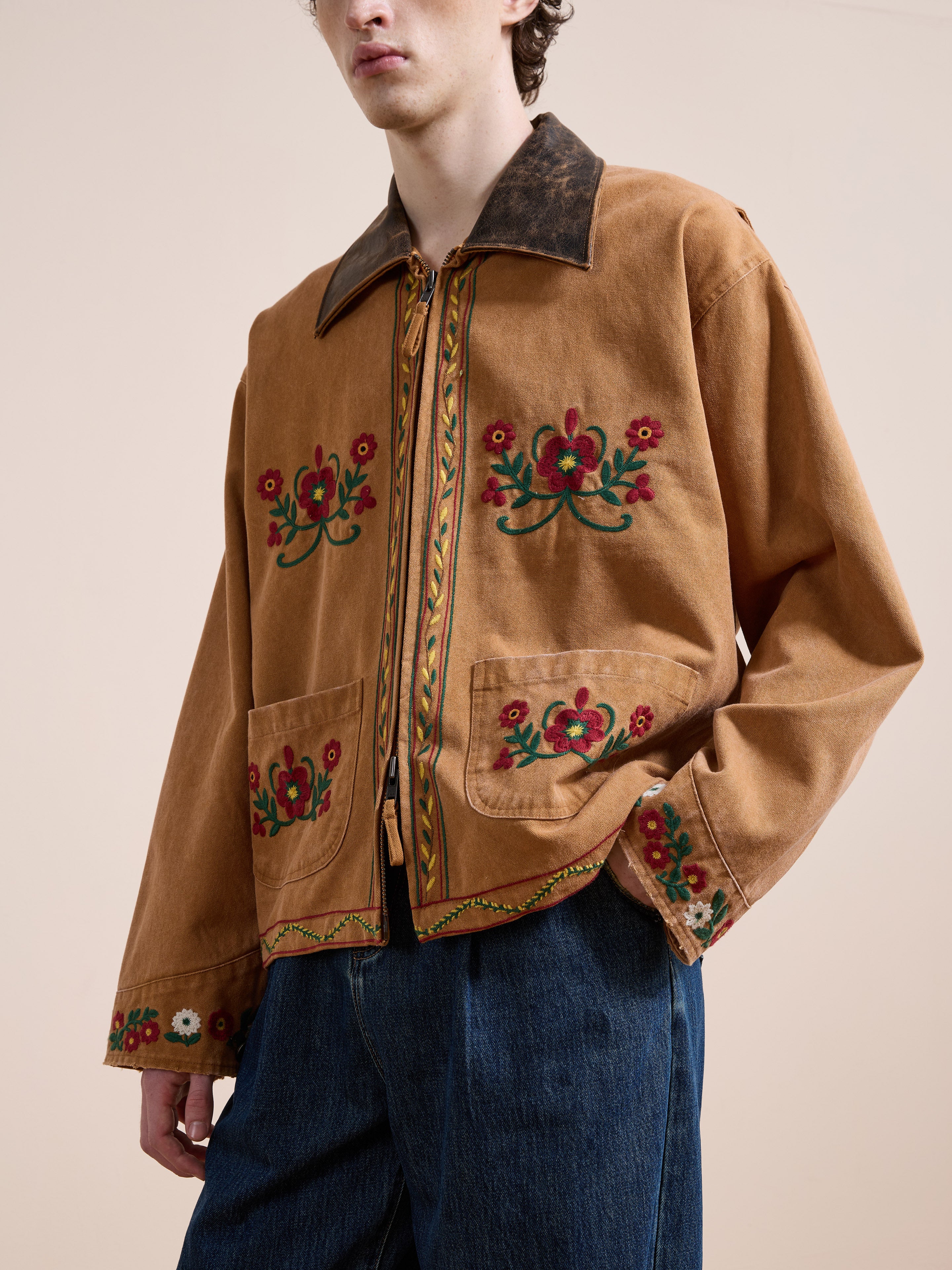A person wearing the Found Rosette Canvas Work Jacket in tan, featuring vibrant floral embroidery on the front, sleeves, and trim, paired with blue jeans against a plain background.