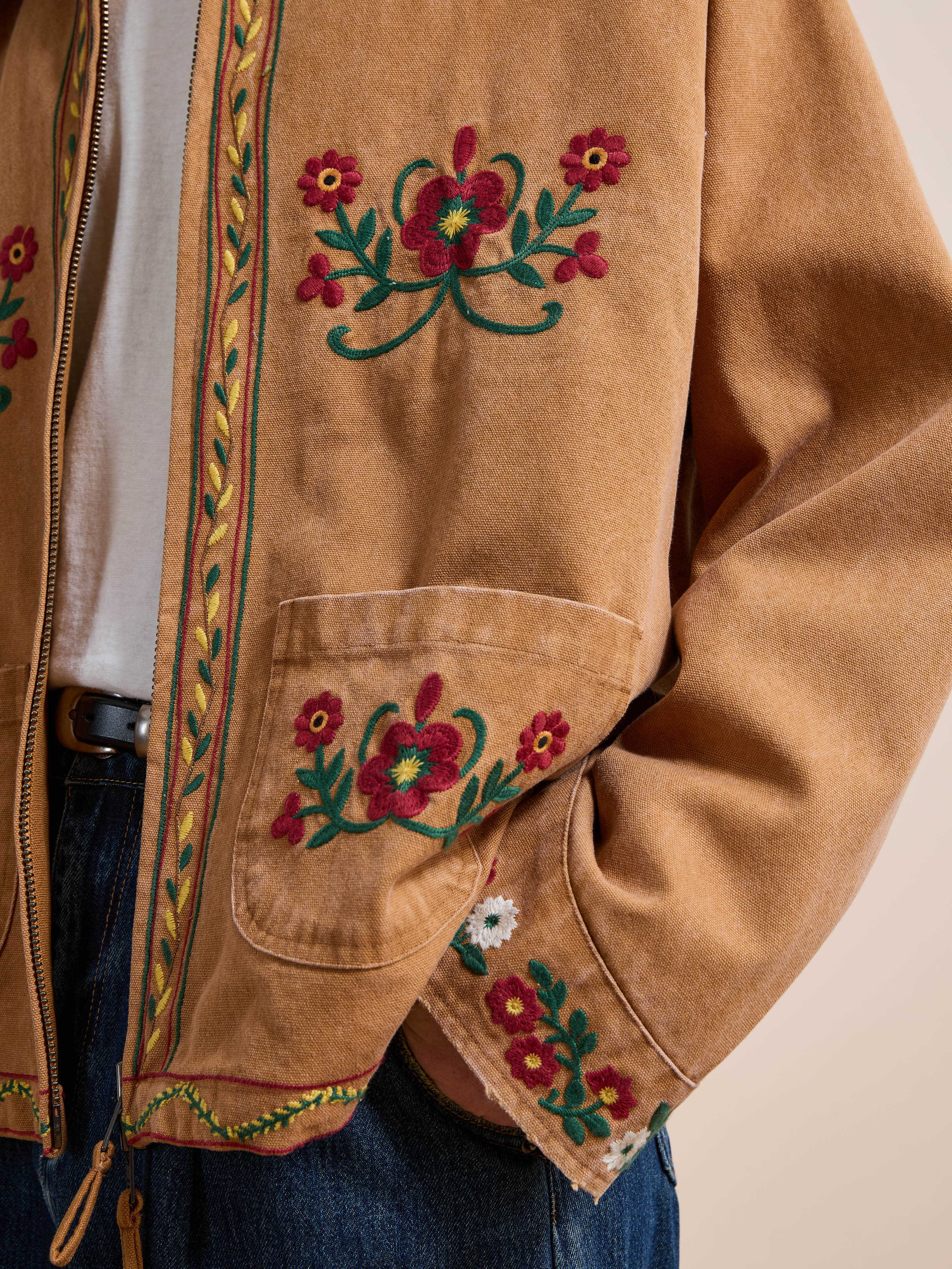 A person wears the Found Rosette Canvas Work Jacket, featuring vintage brown fabric with red and green floral embroidery on the front, pocket, and sleeve, paired with a white shirt and blue jeans.