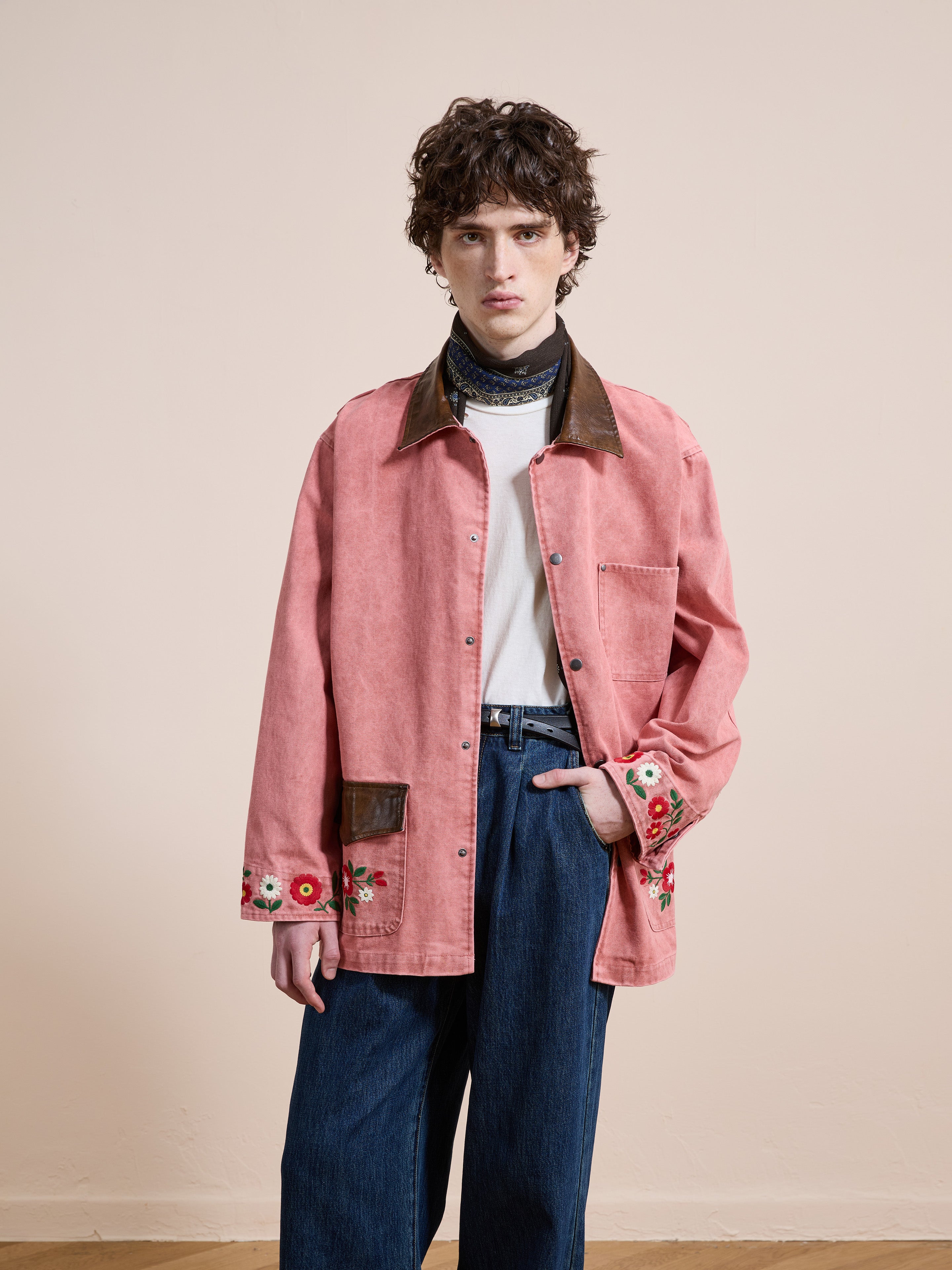 A person wears the Found Canvas Embroidered Barn Jacket, featuring a brown collar and floral embroidery, styled with a white shirt, blue jeans, and a scarf, standing against a plain beige background.