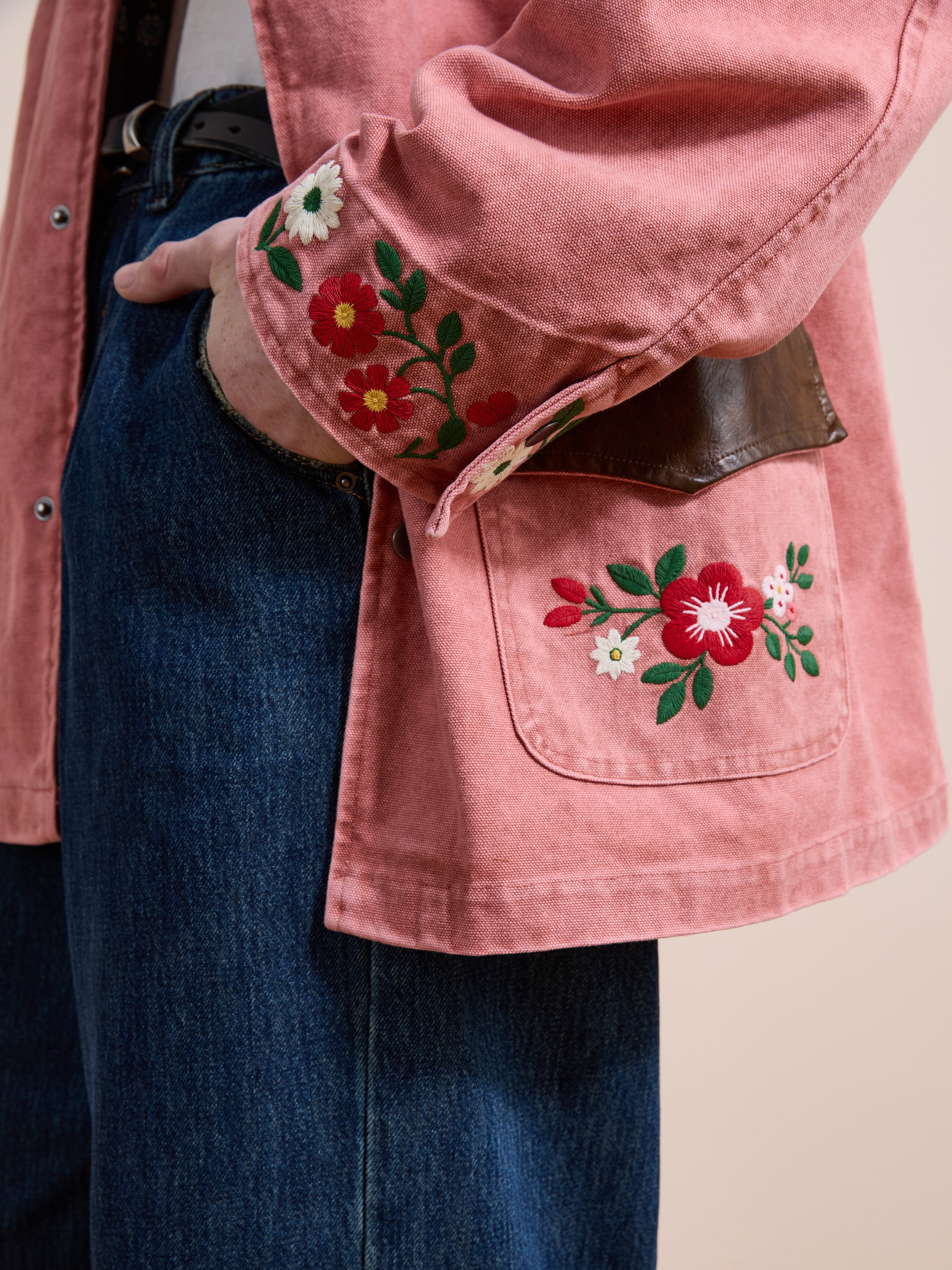 A person wears the Found Canvas Embroidered Barn Jacket featuring pink floral designs on the sleeve and pocket, paired with blue jeans. Their hand is tucked into a pocket.