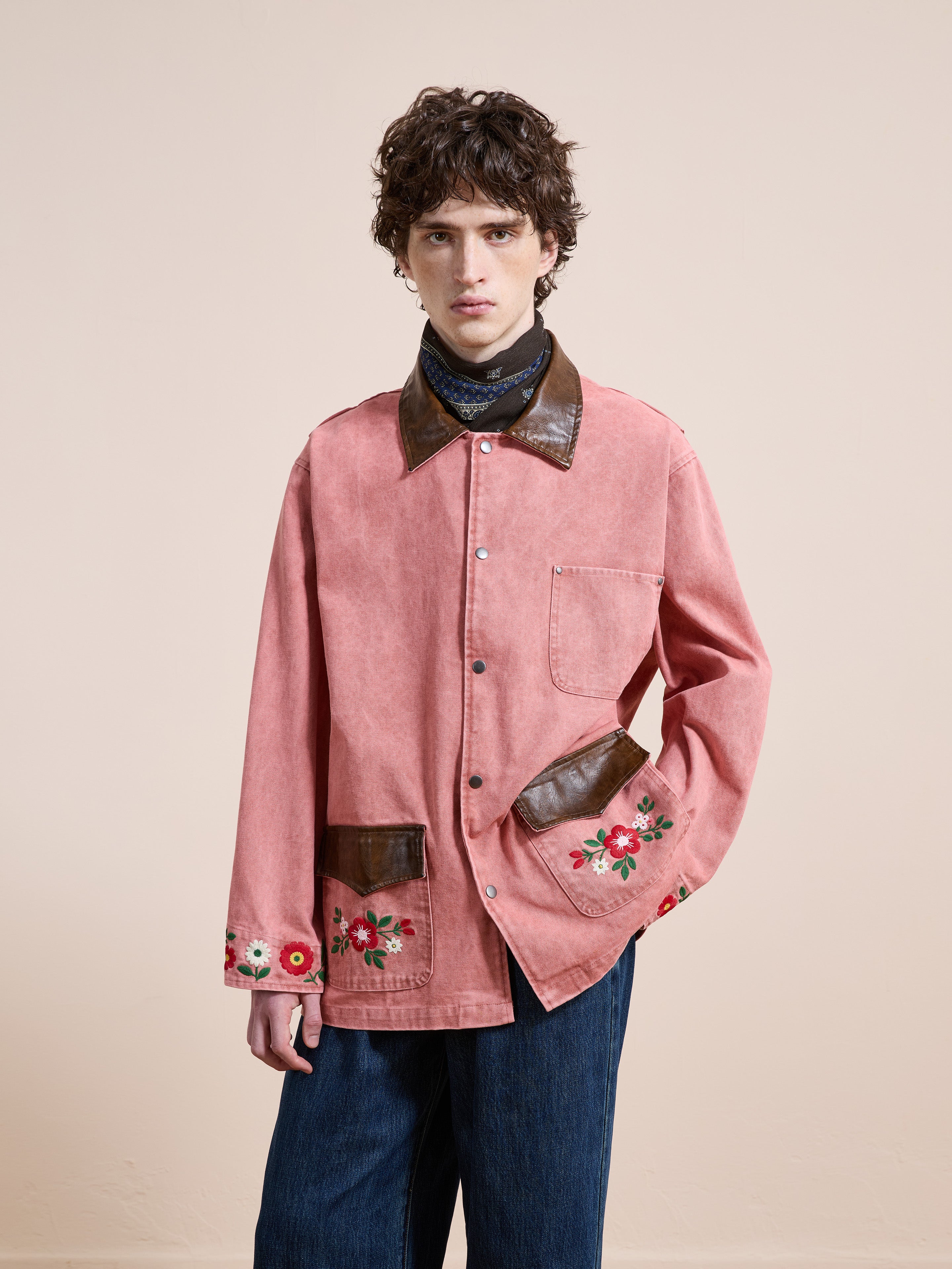 A person models the Found Canvas Embroidered Barn Jacket, featuring pink floral embroidery and brown leather-trimmed pockets and cuffs, styled with blue jeans against a plain beige background.