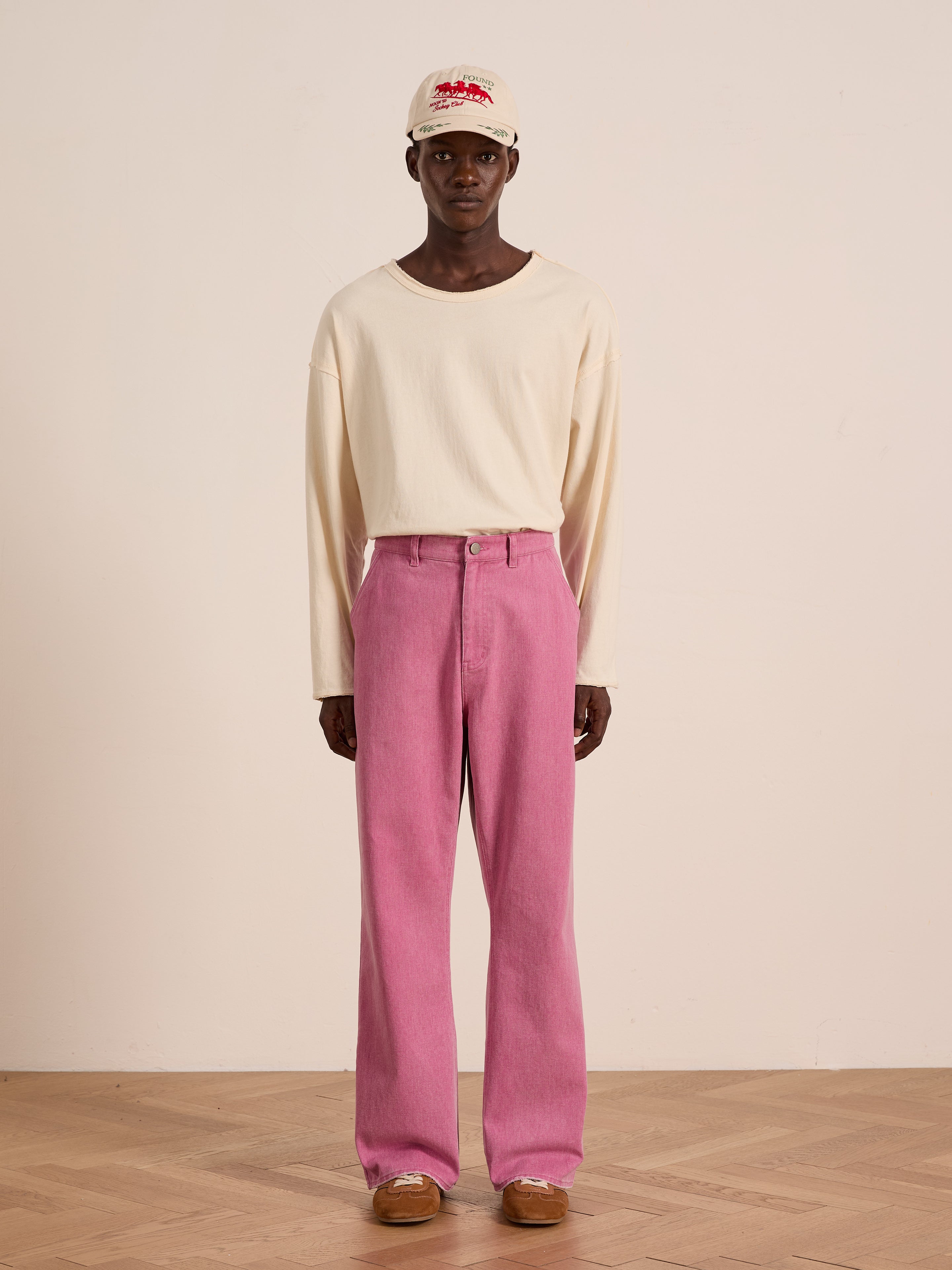 A person indoors models Flare Work Pants by FOUND—styled with a cream long-sleeve shirt, tan shoes, and a light baseball cap for a fresh take on unisex workwear.