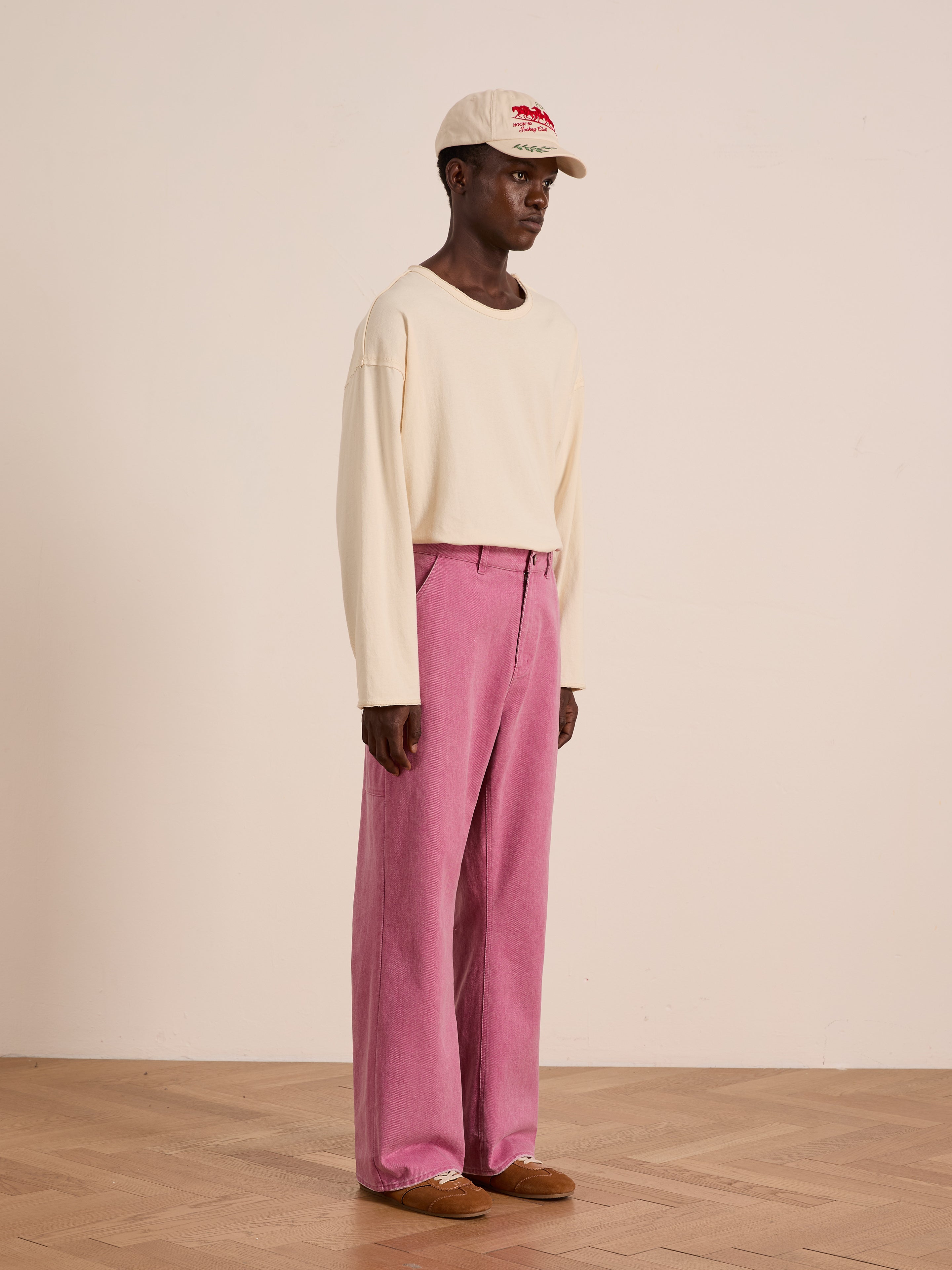 Indoors, a person wears a cream long-sleeve shirt, brown shoes, a beige cap, and FOUND Flare Work Pants in pink. The plain background emphasizes the effortless, unisex workwear style of this look.