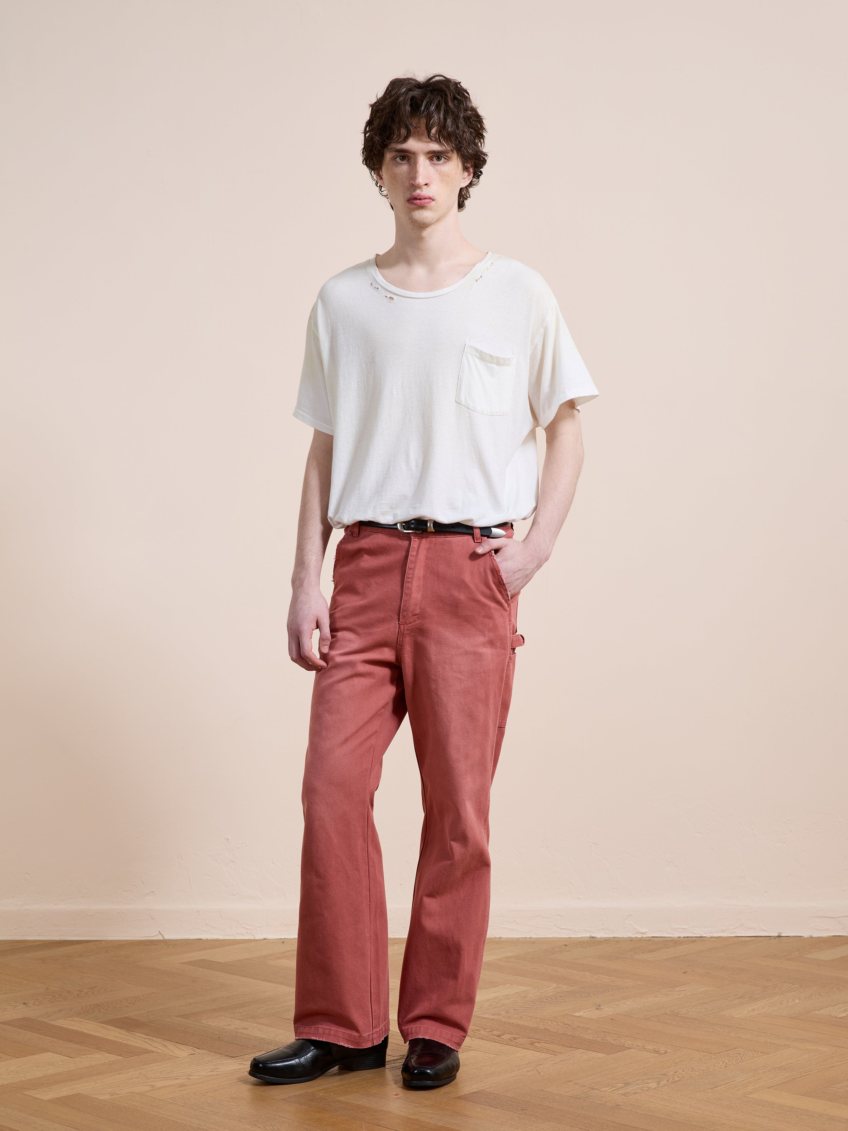 Indoors on a wooden floor, a person faces the camera wearing a white T-shirt, FOUND Canvas Faded Pants with a black belt, and black shoes, one hand in their pocket.