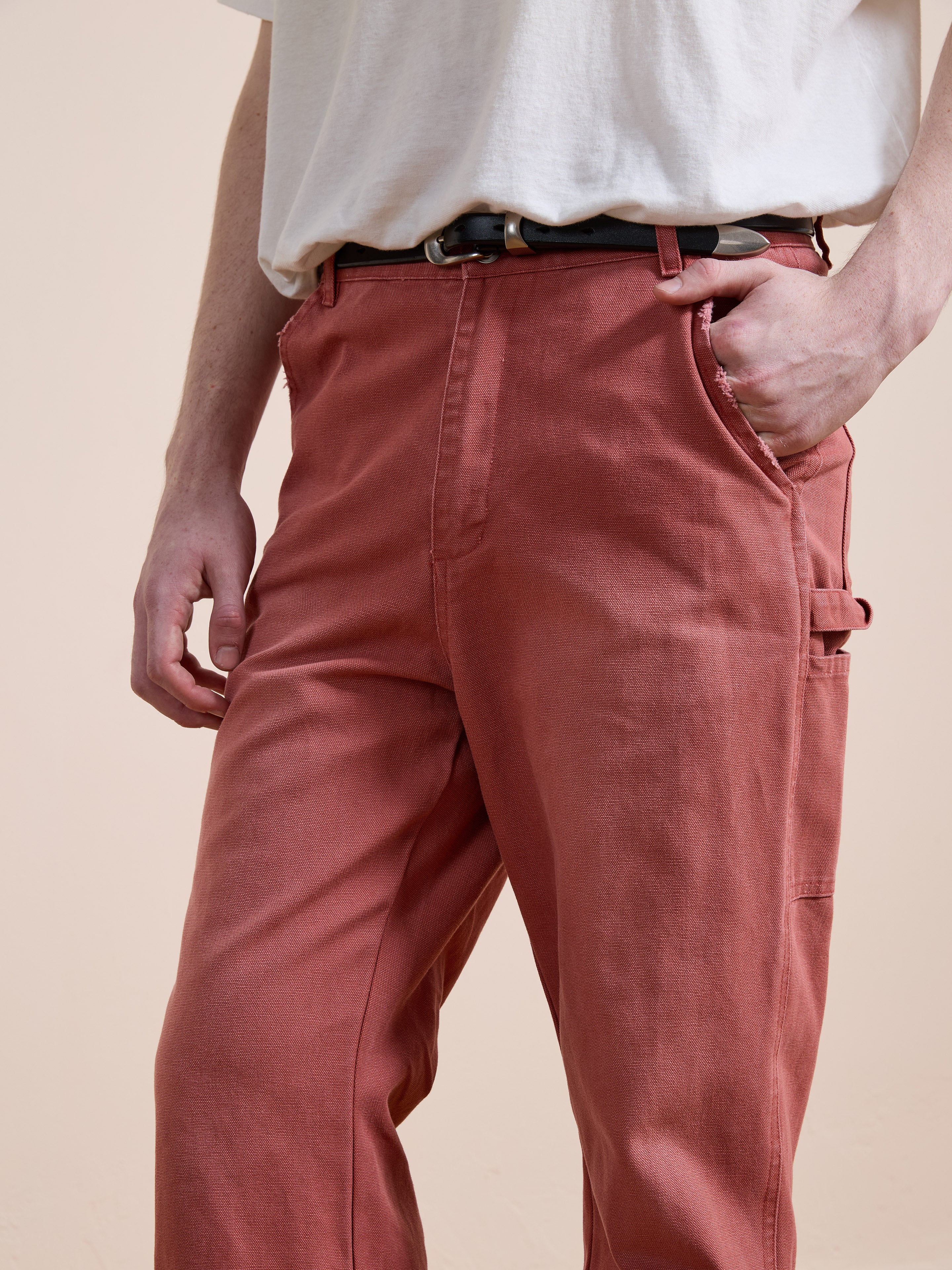 Model wears FOUND’s Canvas Faded Pants in vintage salmon wash—high-waisted, loose-fitting style—paired with a white t-shirt and black belt, hand in pocket, against a plain background.