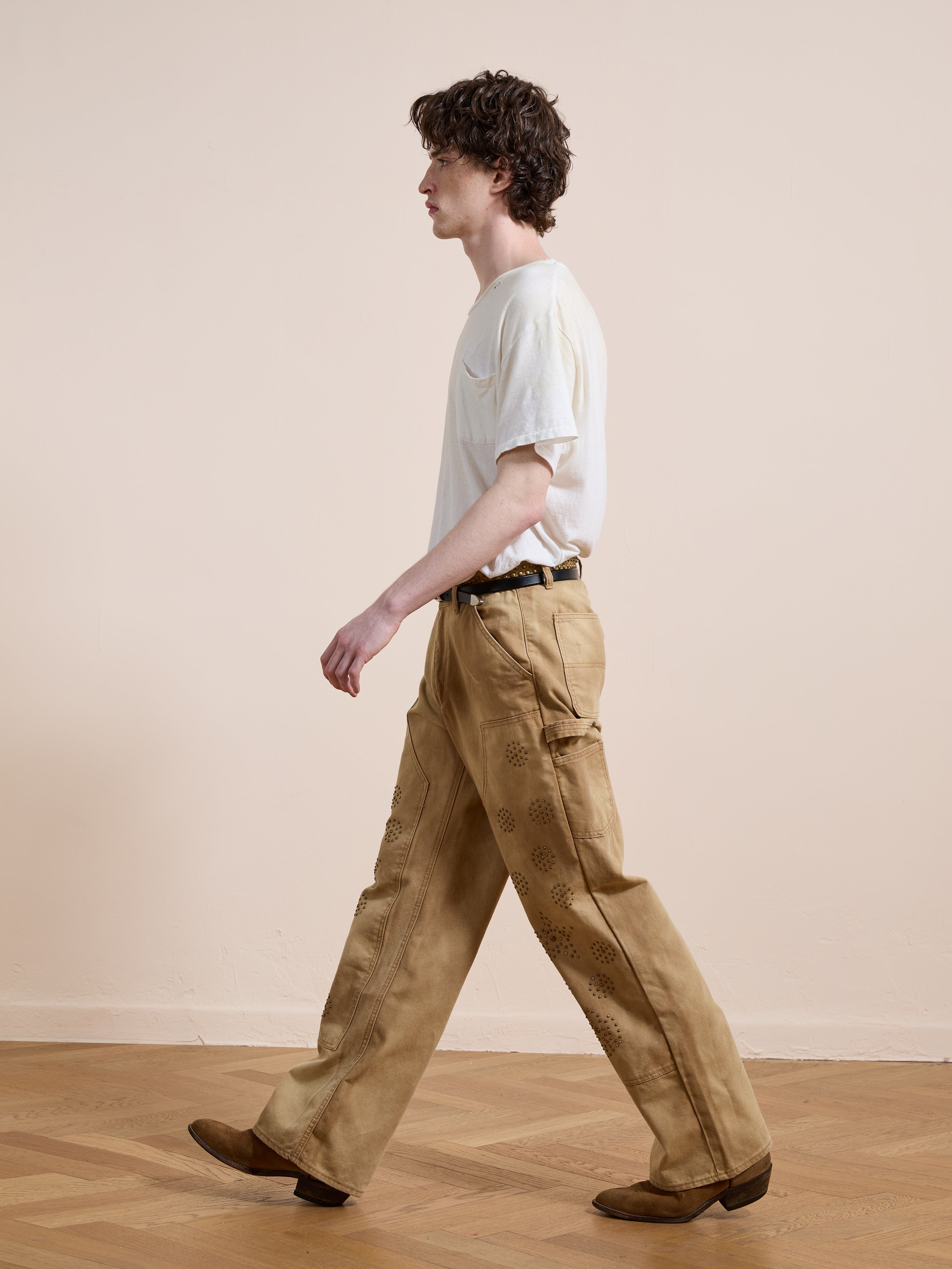 A person with curly hair walks indoors on a wooden floor, wearing a white t-shirt, brown shoes, and FOUND Canvas Studded Work Pants—unisex, light brown, workwear-inspired wide-leg pants with patterns.