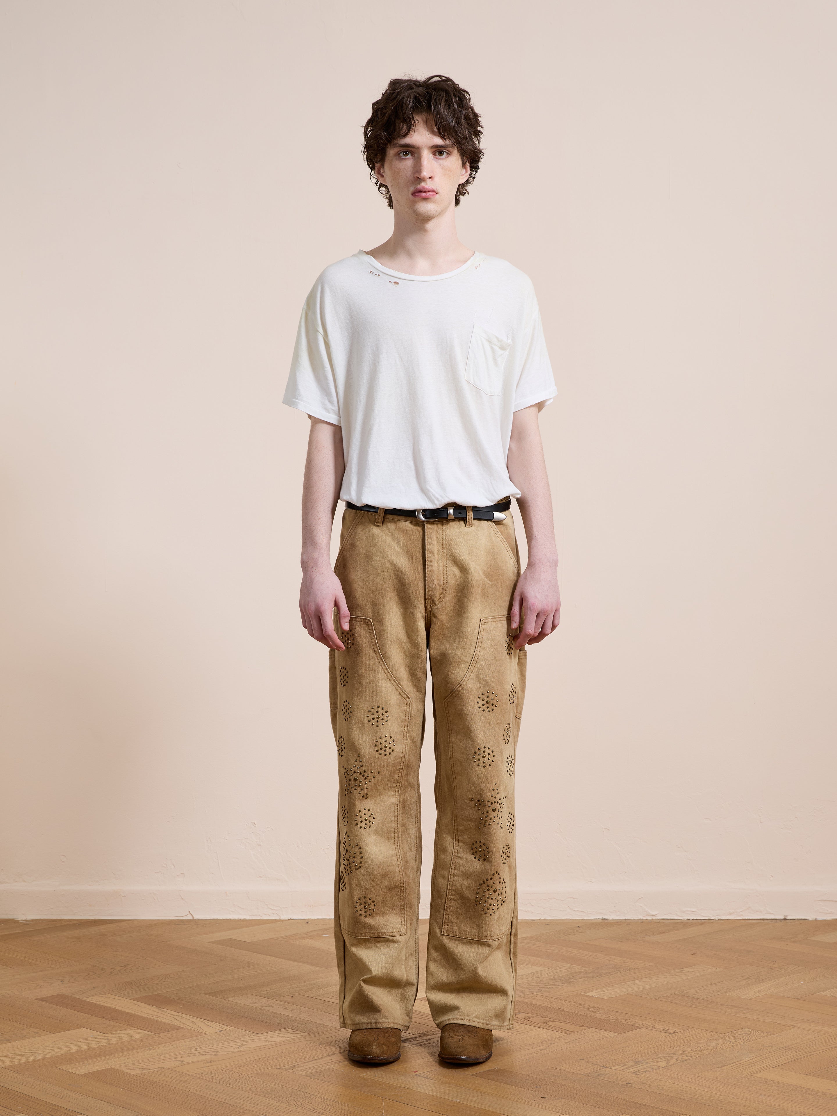 Person stands indoors against a plain wall, wearing a white t-shirt, FOUND Canvas Studded Work Pants with a tan pattern, a black belt, and brown shoes.