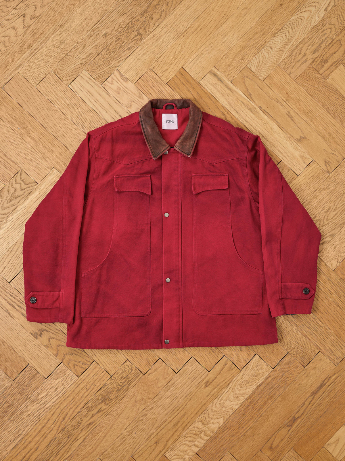 The Canvas Utility Barn Jacket by Found, featuring a red suede finish, a corduroy collar, buttoned cuffs, two chest pockets, and snap buttons, is laid flat on a herringbone wood floor.